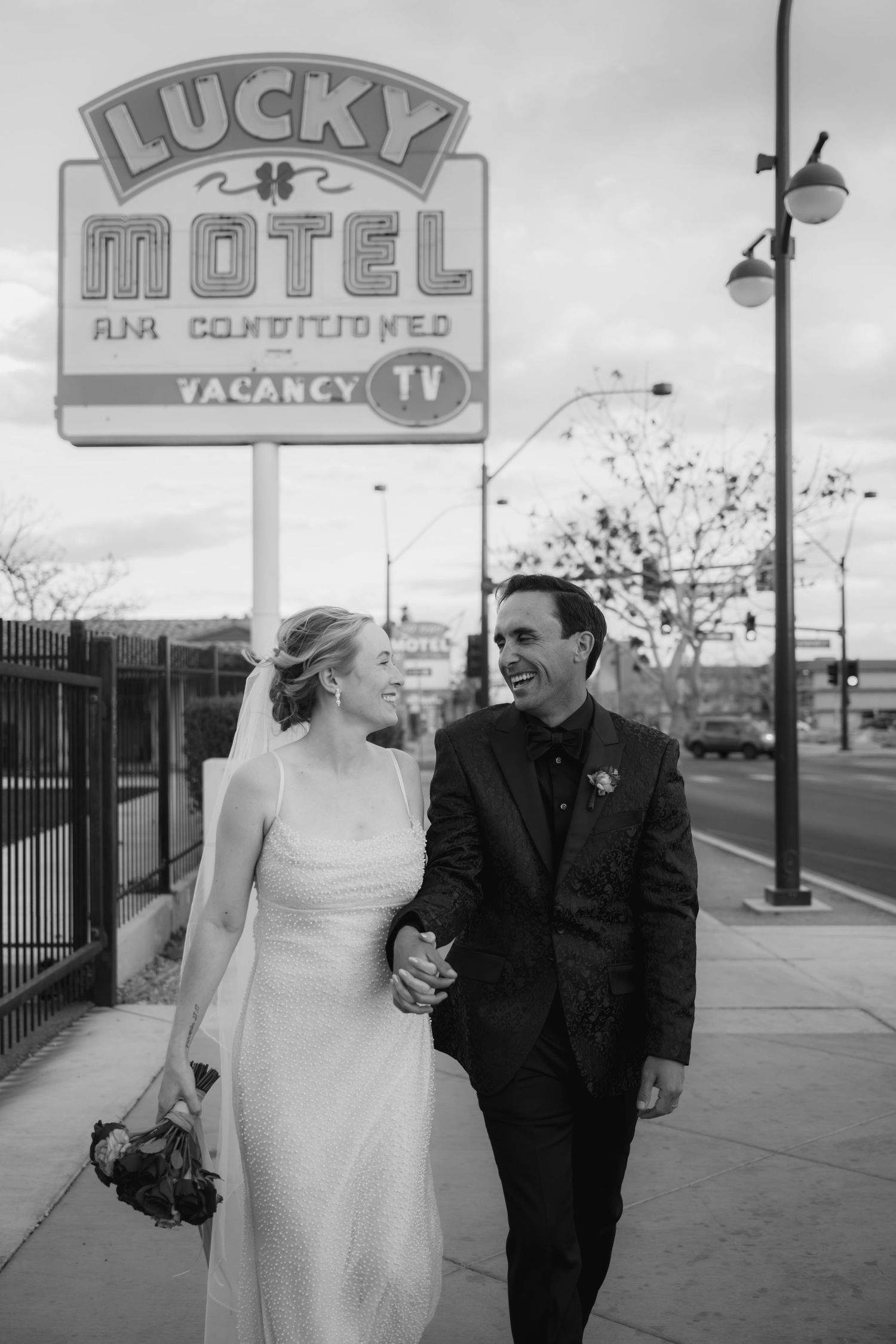 Black and white photo of a smiling bride and groom holding hands in front of the Lucky Motel sign in Las Vegas.