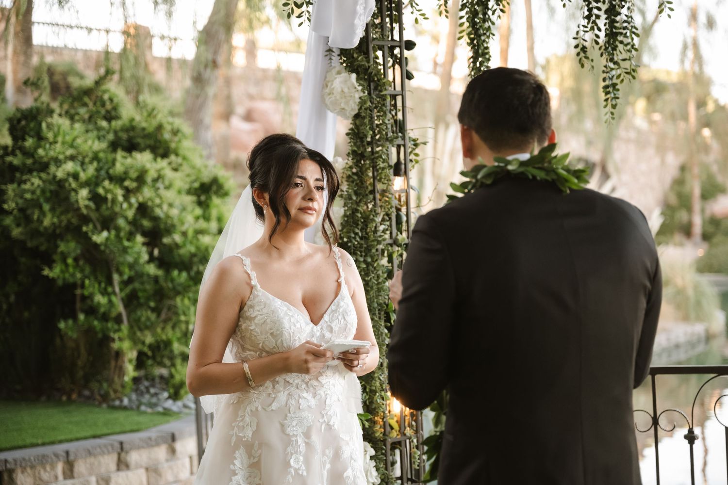 Bride in lace gown reading vows to groom under floral arch at outdoor wedding ceremony.