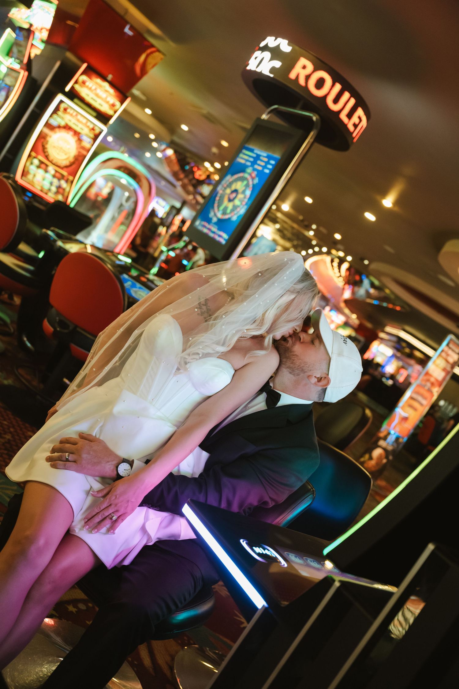 A couple in formal attire dances together at a dimly lit venue with colorful lighting and bar decor in the background.