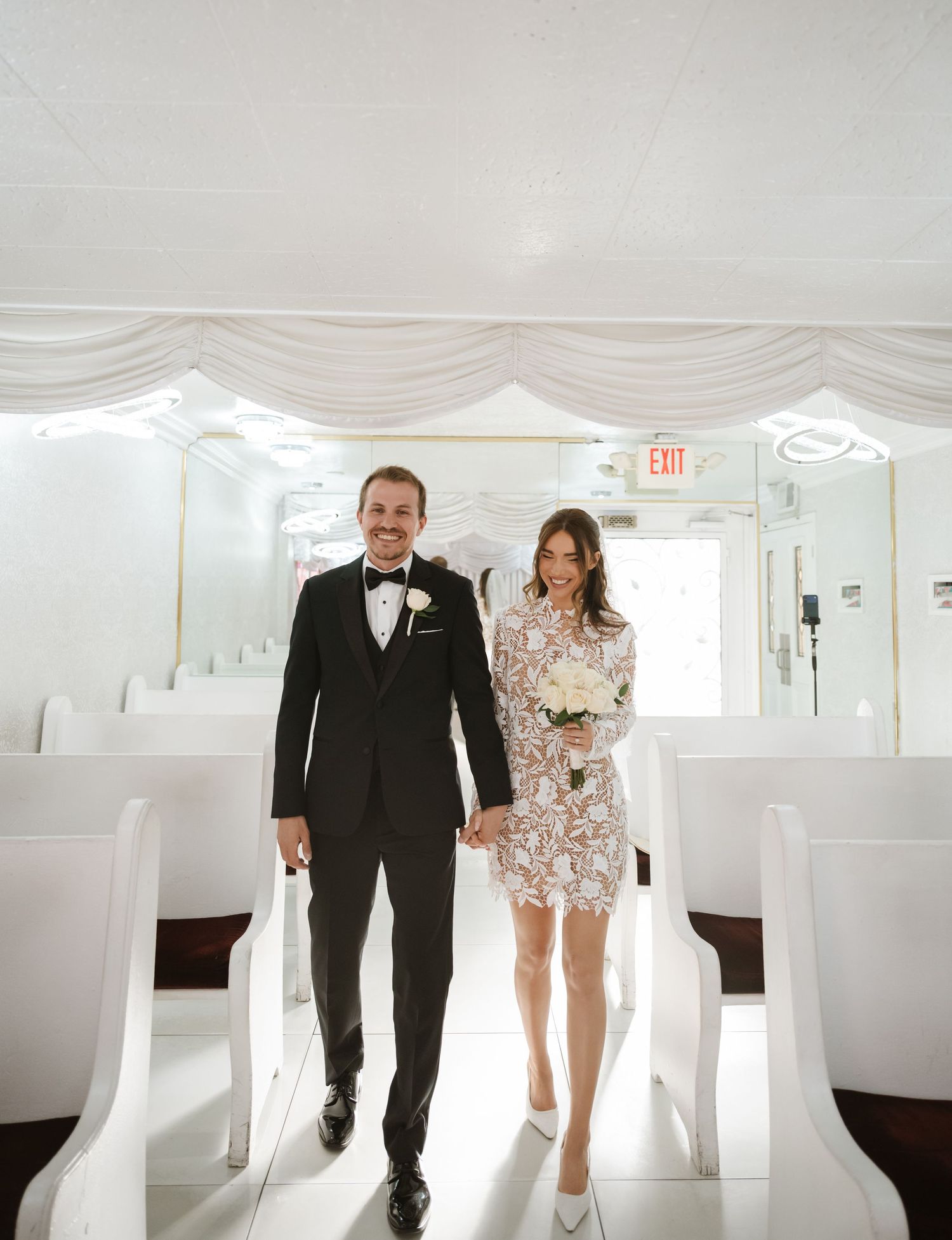 Bride in lace mini dress and groom in black tuxedo walking down white chapel aisle after wedding ceremony.