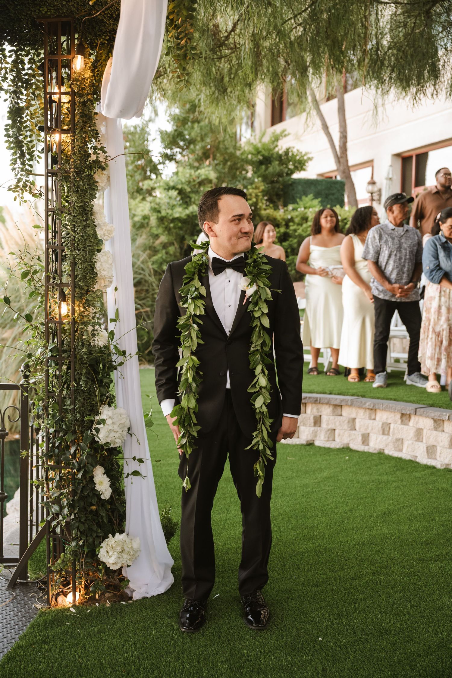Smiling groom in black tuxedo and green lei stands at outdoor wedding ceremony altar.