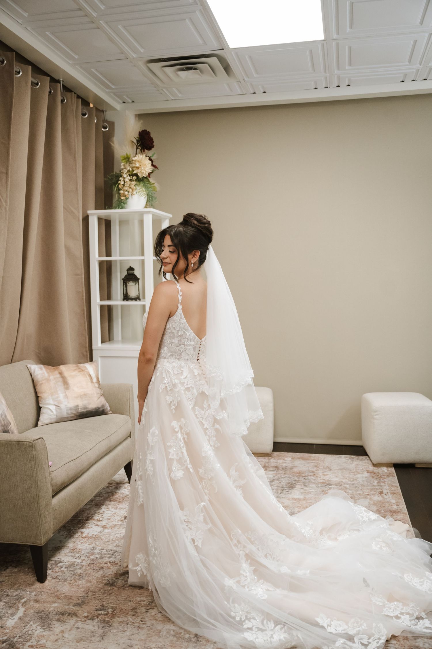 Bride in a lace wedding gown with veil and train stands in an elegant bridal suite, viewed from behind.