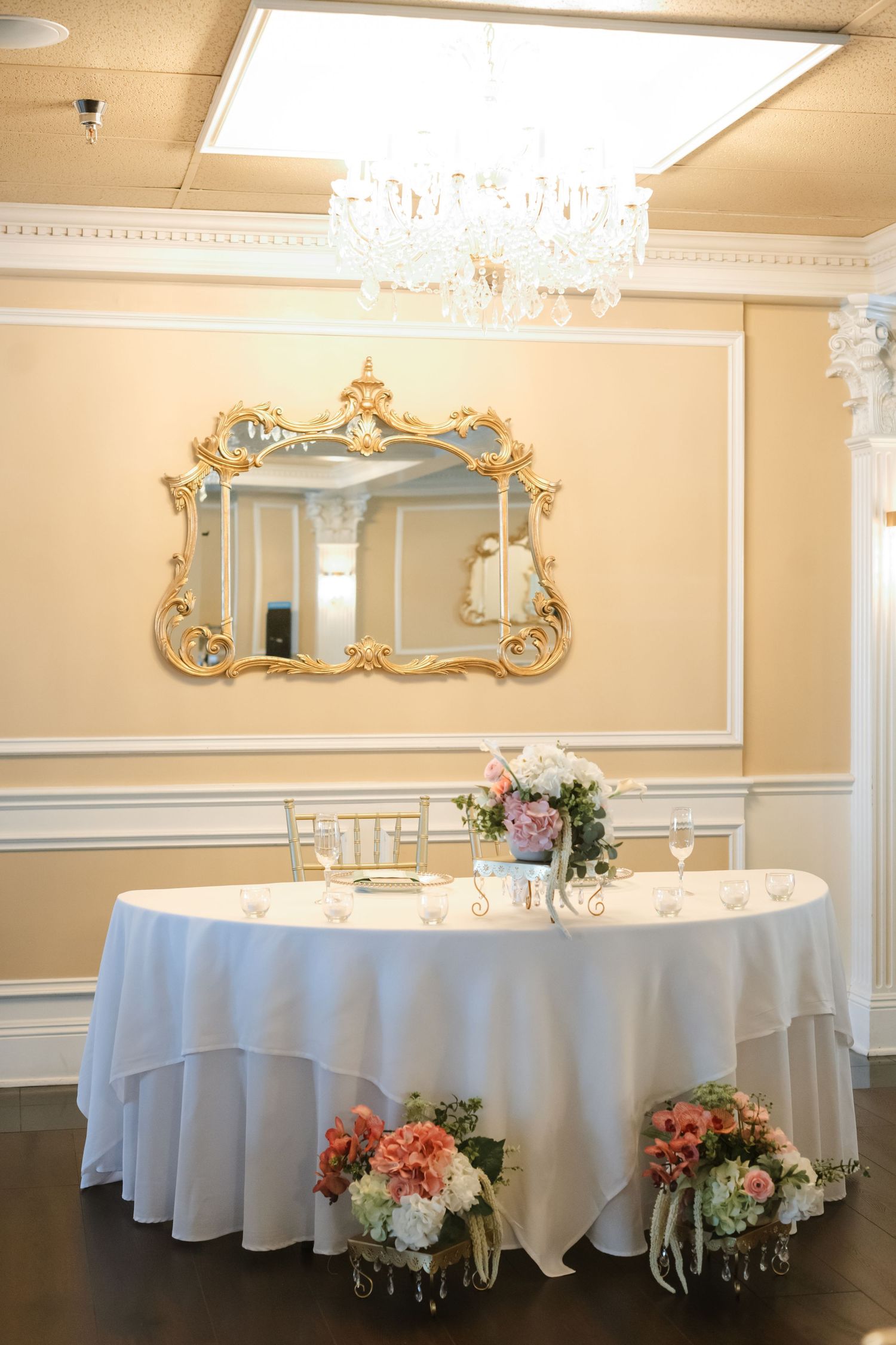 Elegant wedding sweetheart table with white linen, floral arrangements, gold mirror, and crystal chandelier.