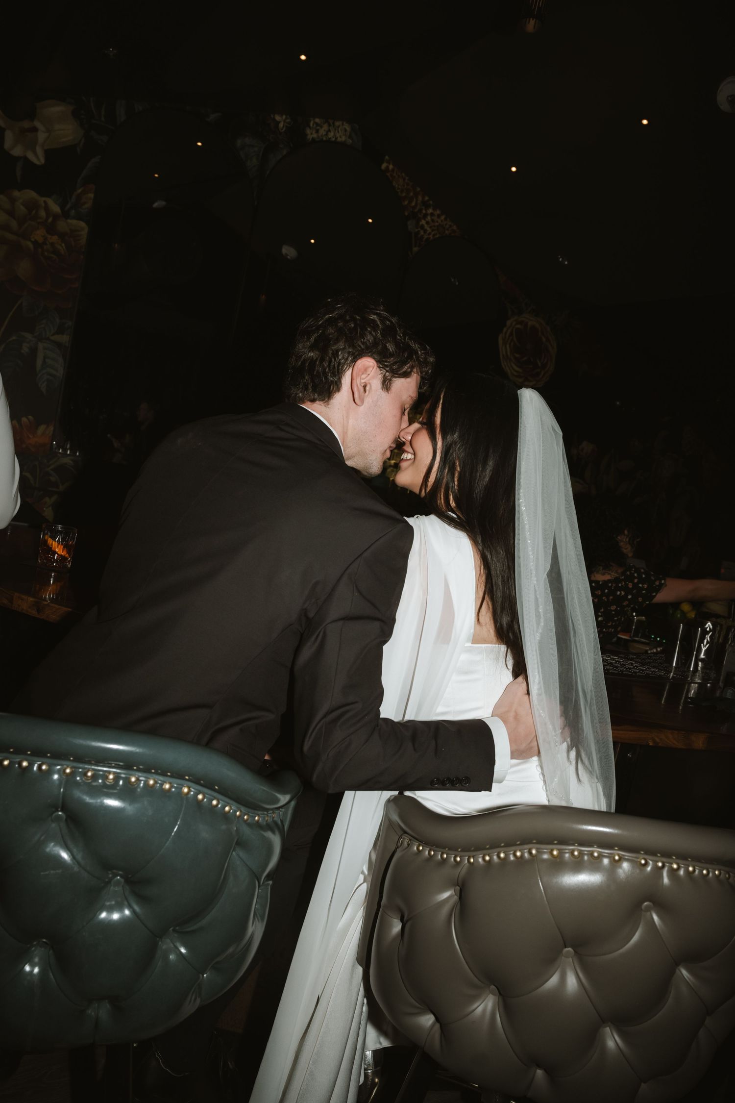 A bride and groom share a romantic kiss at their wedding reception, seated in tufted gray velvet chairs.