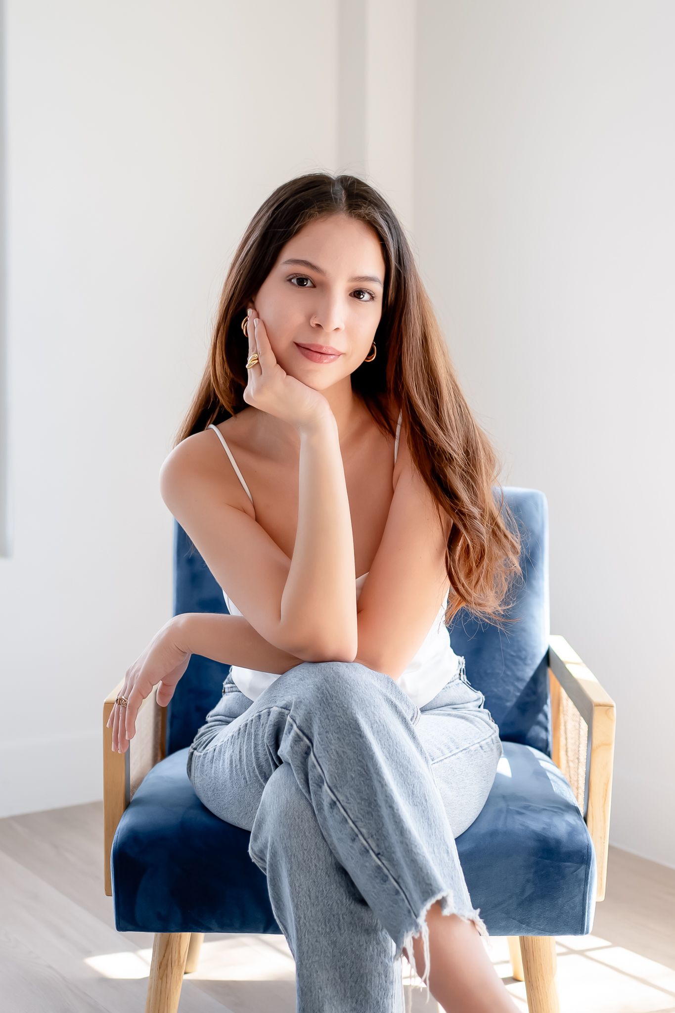 Lifestyle portrait of woman in white top and jeans sitting in blue velvet chair.