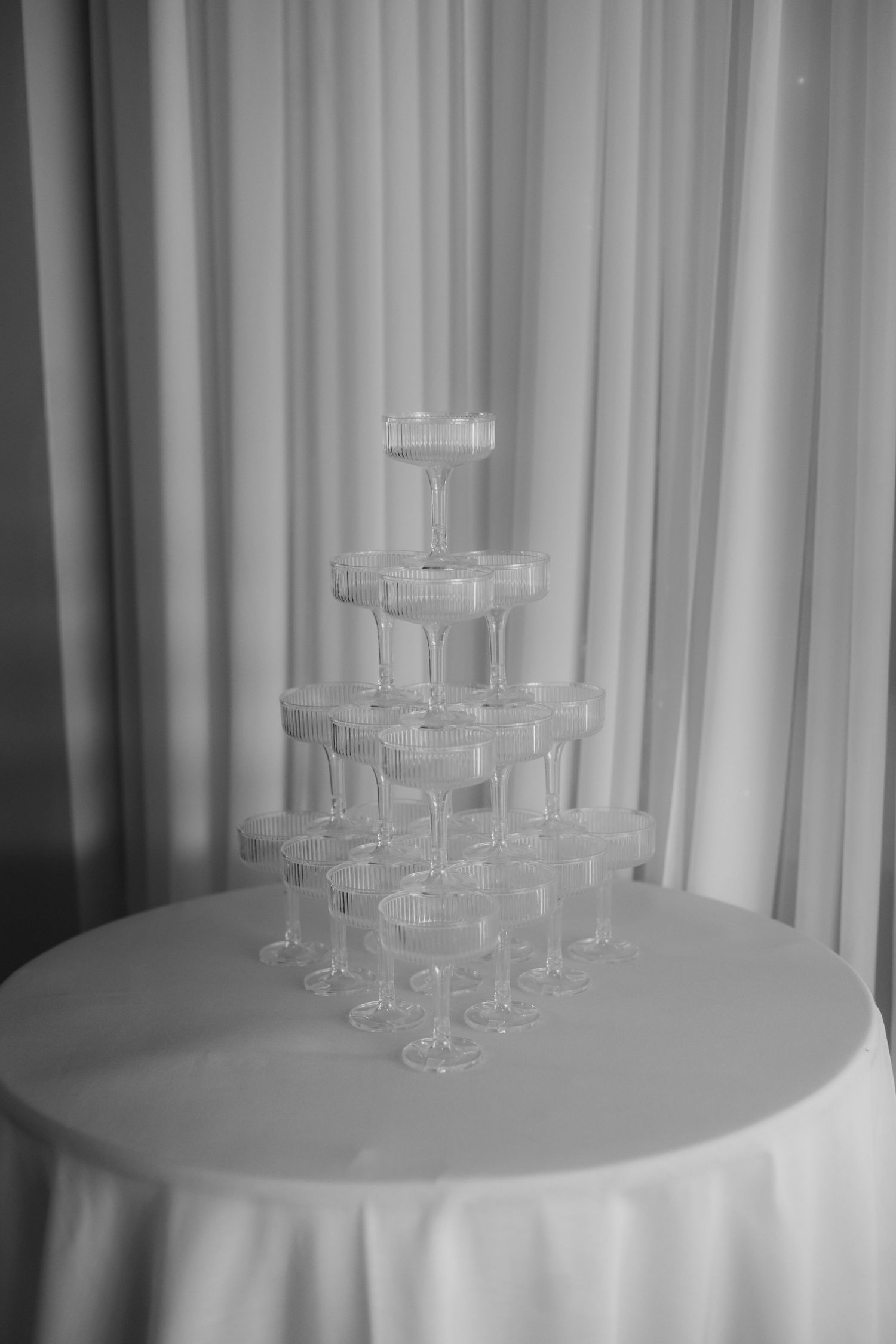 Black and white photo of elegant champagne tower with empty glasses stacked in pyramid on white draped table.