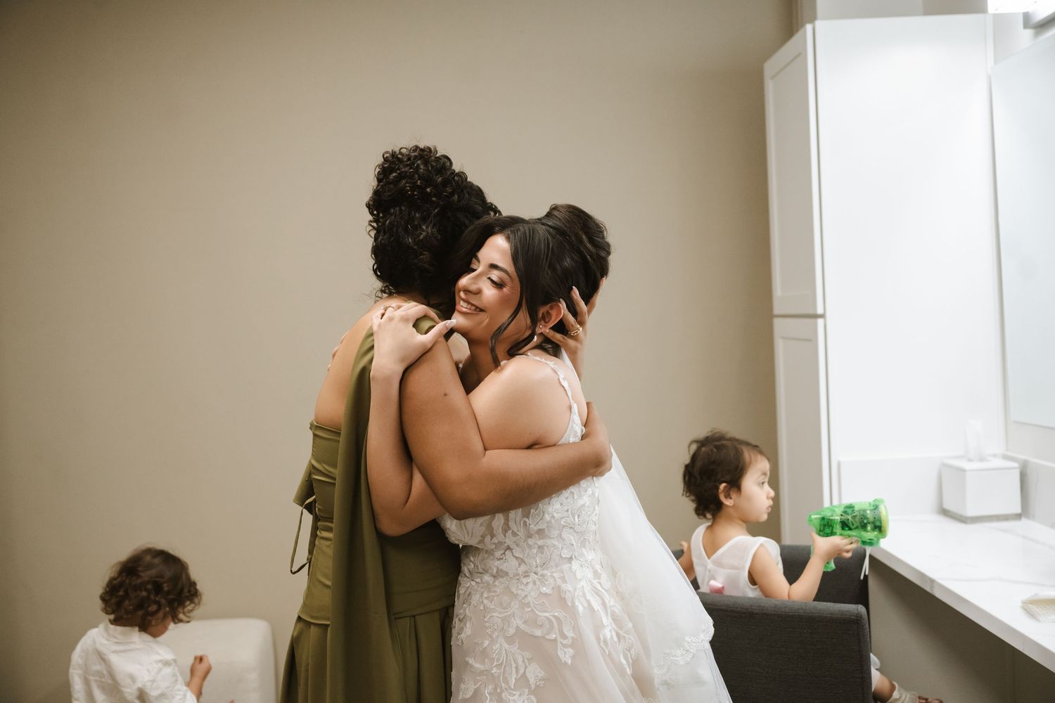 Bride in white lace gown embraces bridesmaid in olive dress, sharing emotional hug before wedding ceremony.