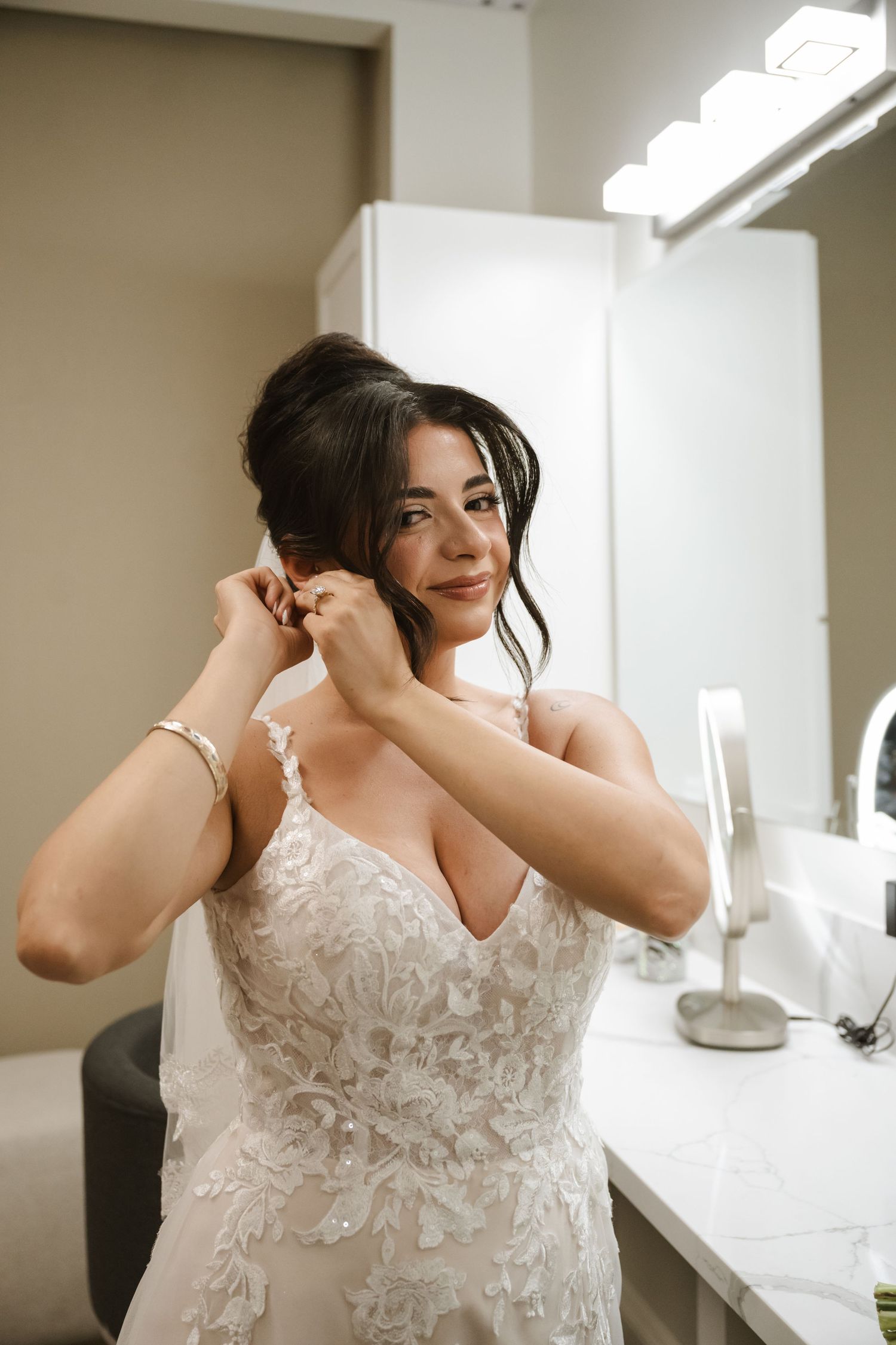 Bride in lace wedding gown putting on earrings at vanity mirror with Hollywood lights.