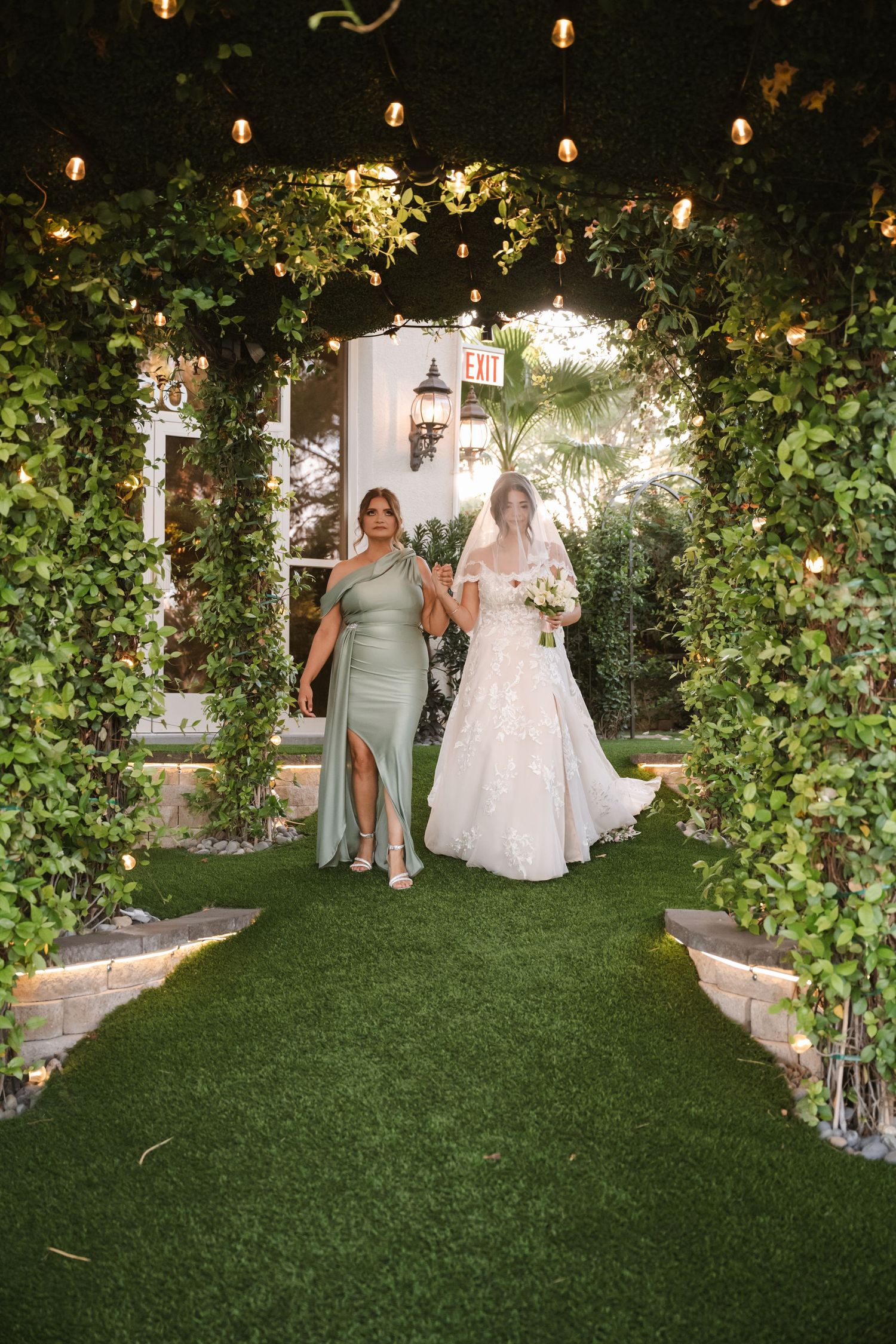 Bride in lace gown walks down ivy-lined aisle with bridesmaid in sage green dress at night.