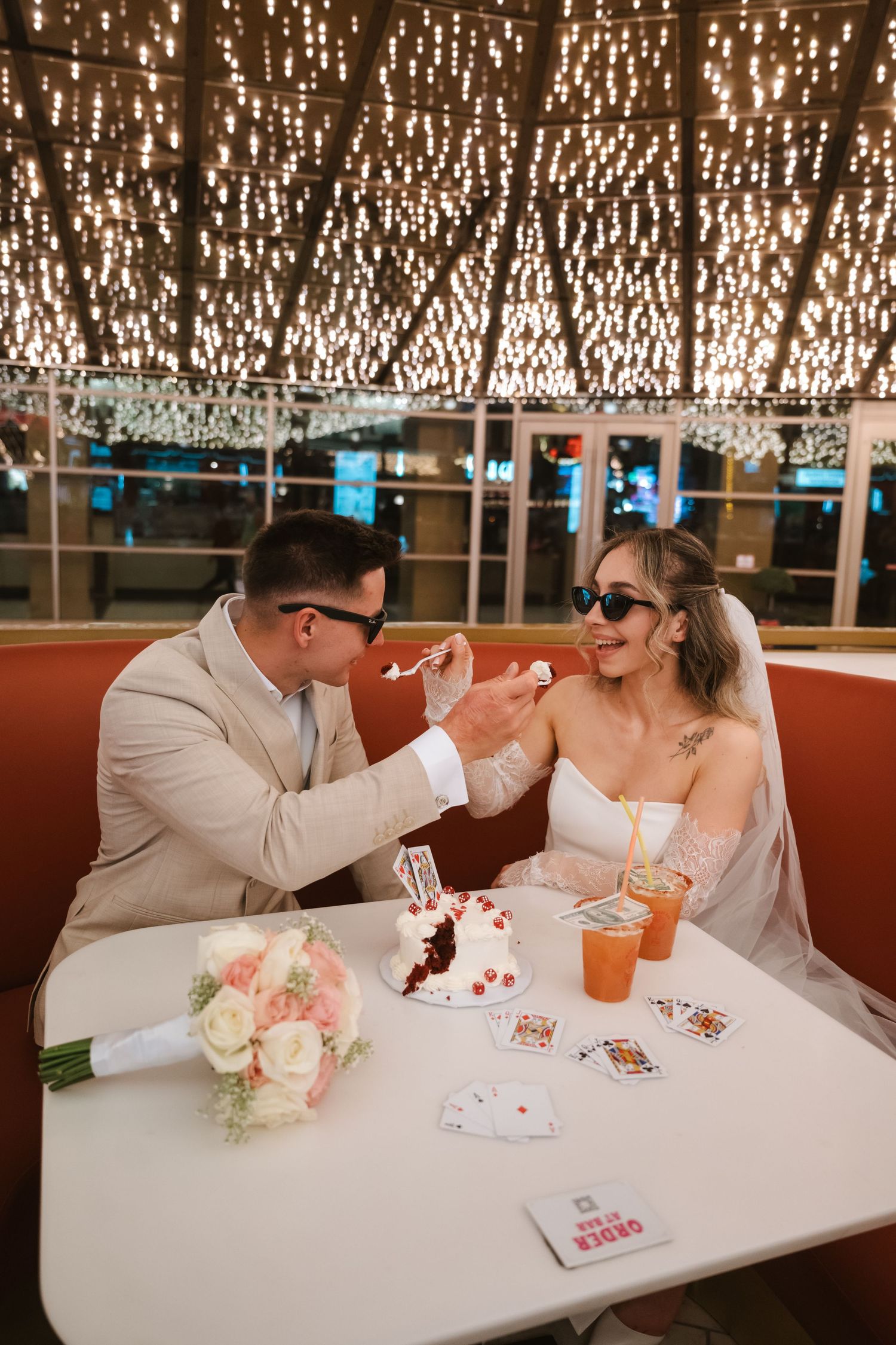 Bride and groom feeding each other cake at a Las Vegas wedding reception table with playing cards and floral bouquet.