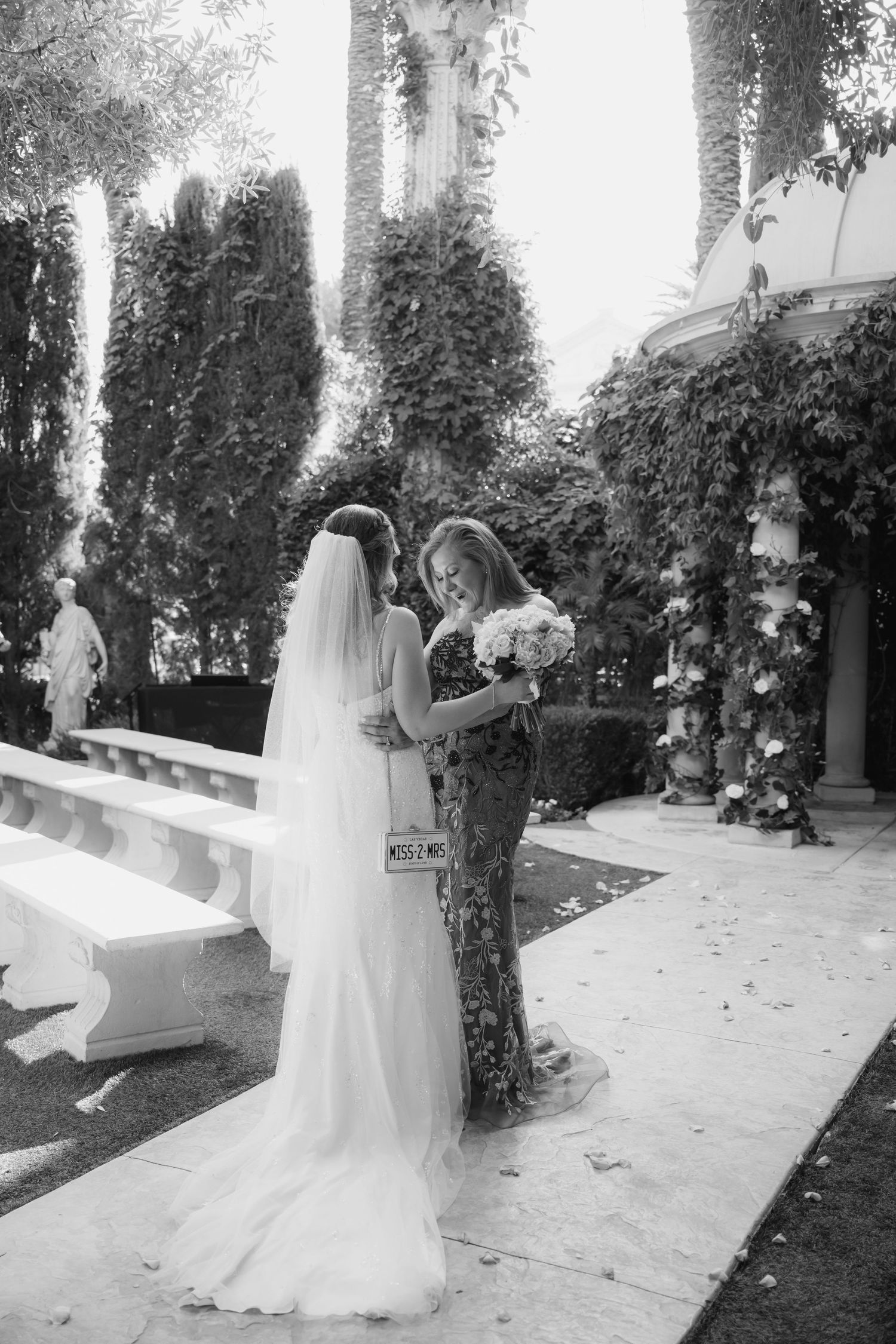 A black and white photo of a bride and guest sharing a moment in a garden setting.