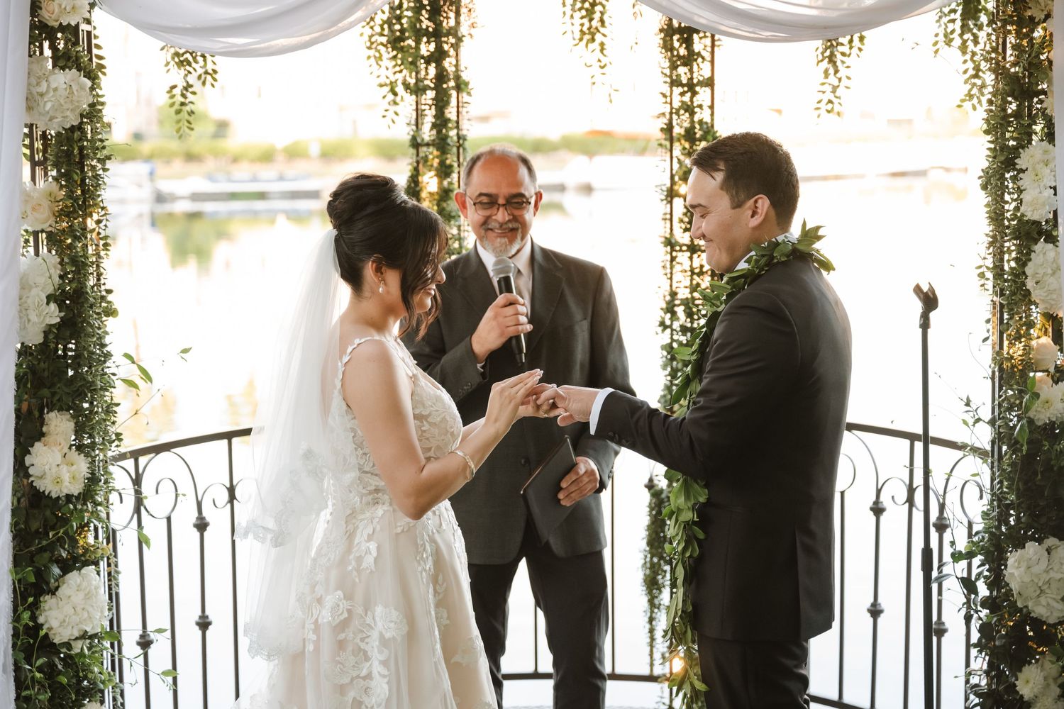 Officiant presides as bride and groom exchange rings at waterfront outdoor wedding ceremony.