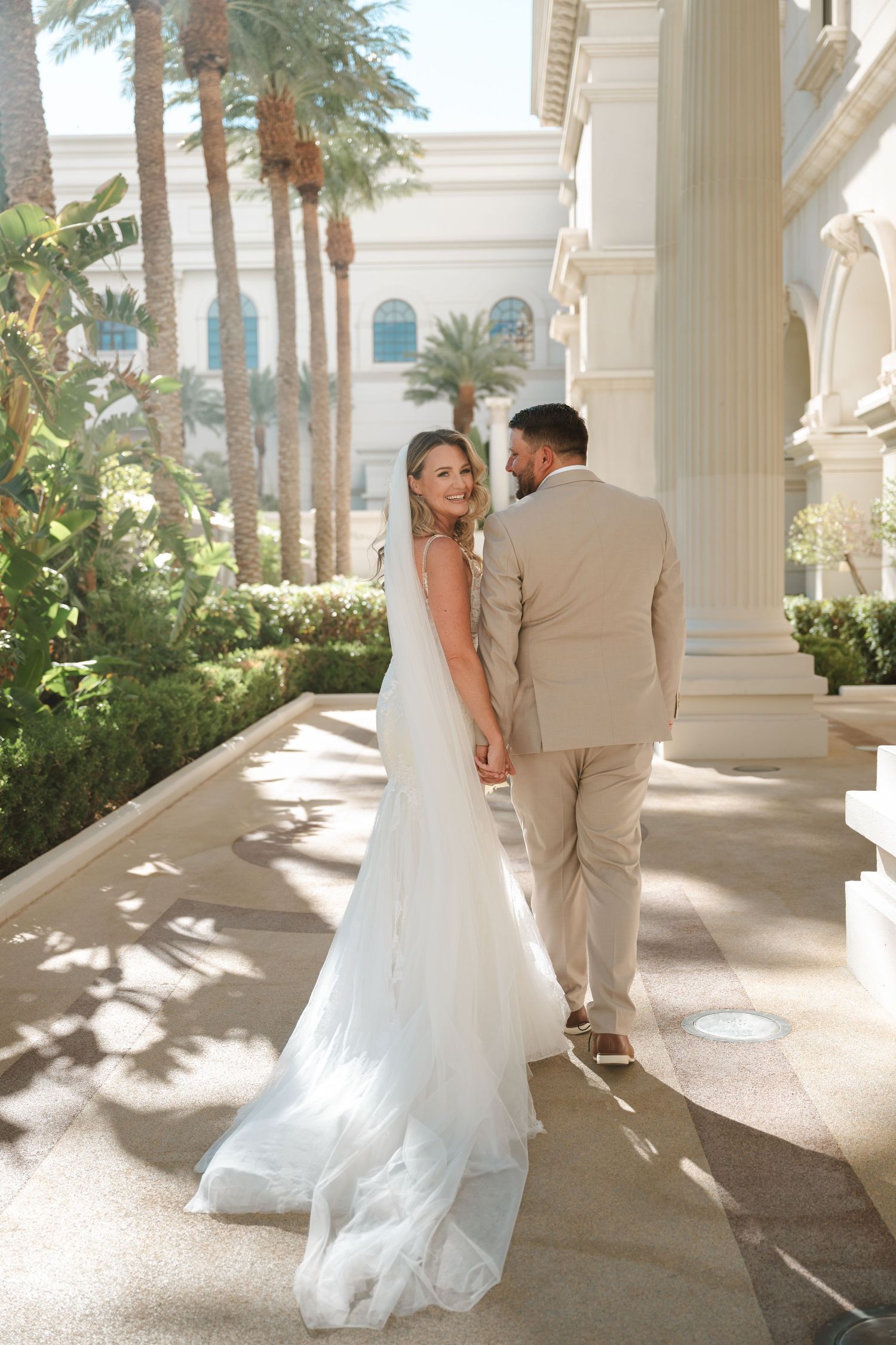 Bride in white gown and groom in beige suit walk hand-in-hand outside a elegant venue with palm trees.