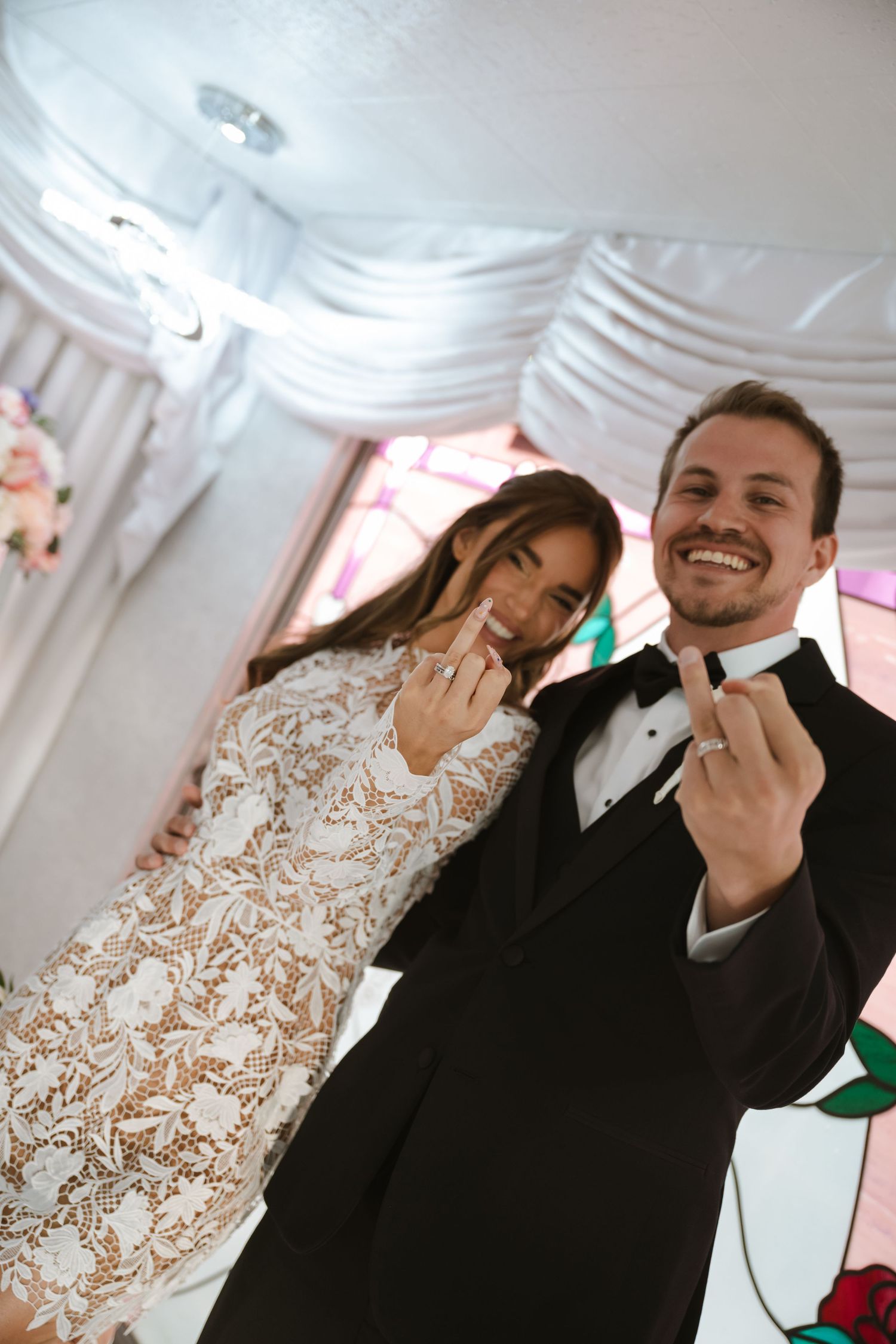 Newlyweds showing off wedding rings at Las Vegas chapel, groom in tuxedo and bride in short lace dress.