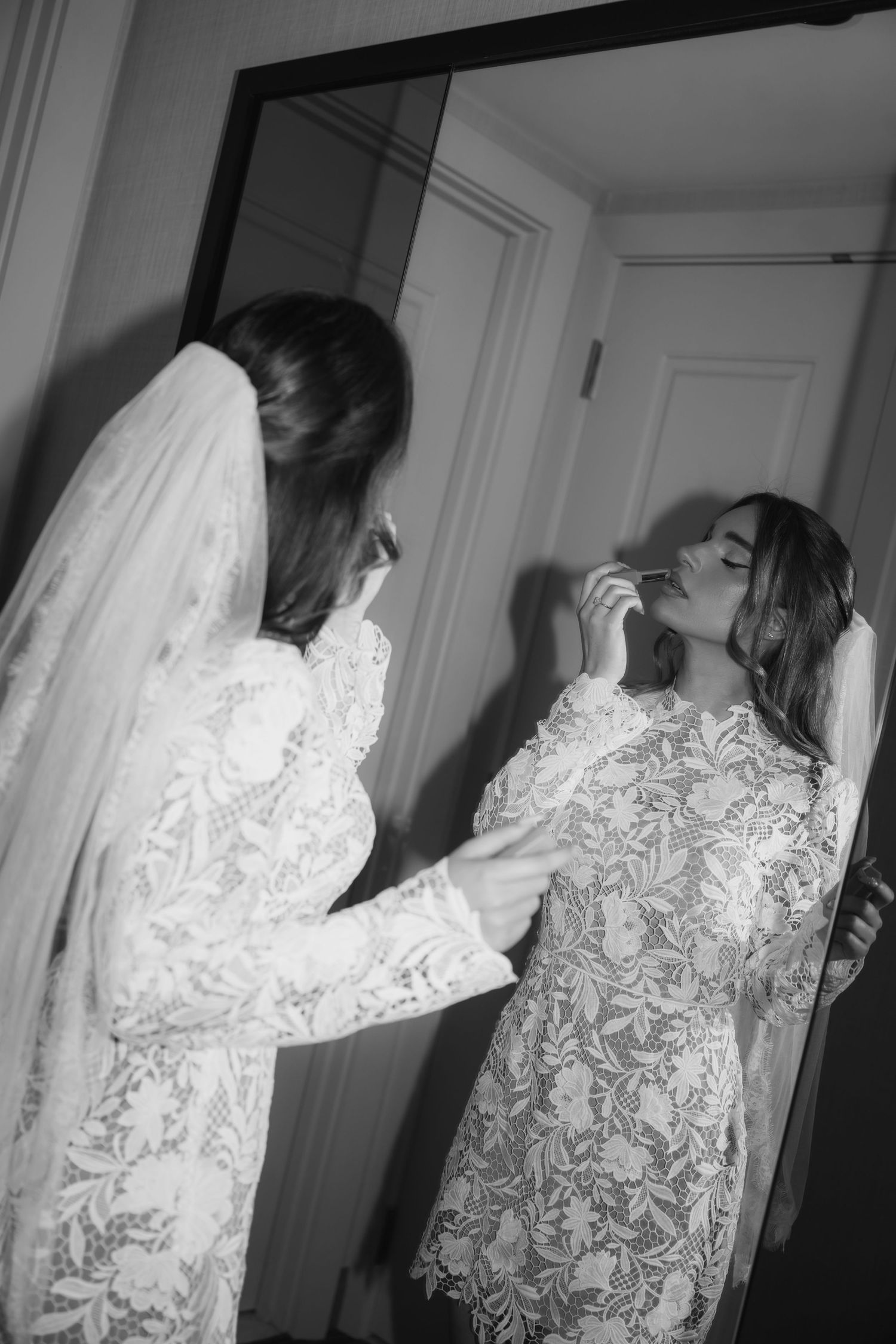 Bride in lace wedding dress and veil applying makeup in mirror, black and white wedding preparation photo.