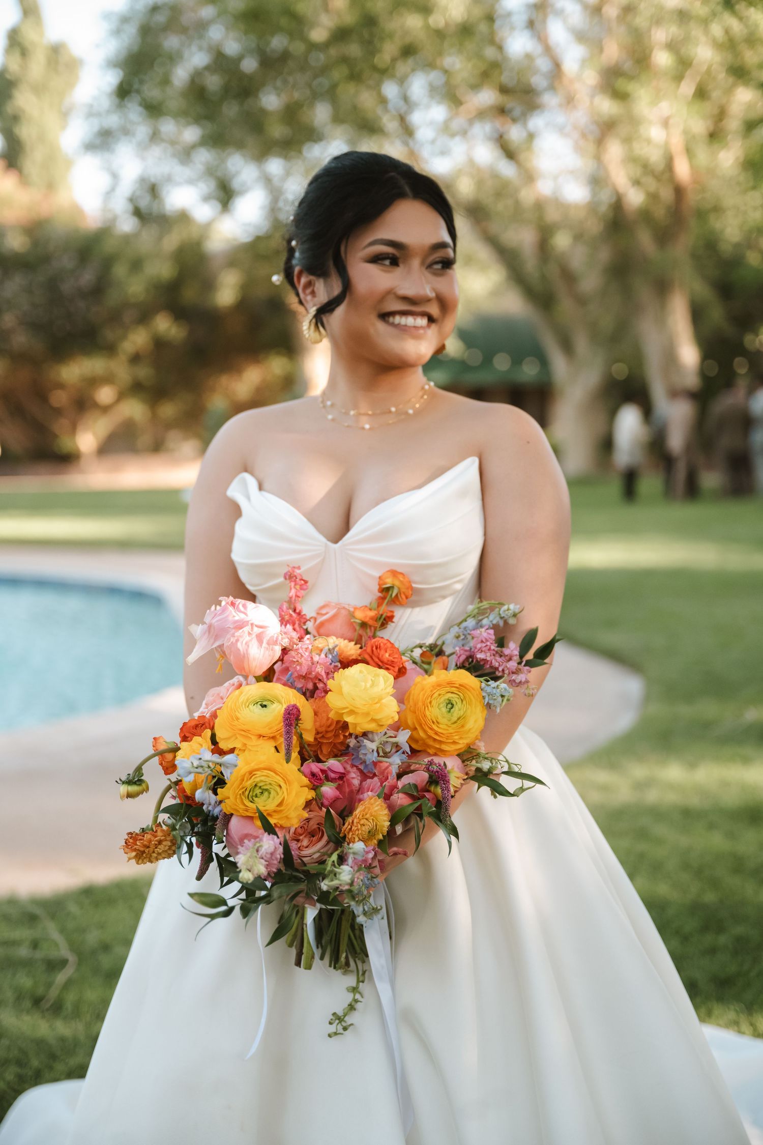 Smiling bride in strapless white gown holds vibrant bouquet of yellow and pink flowers outdoors near a pool.