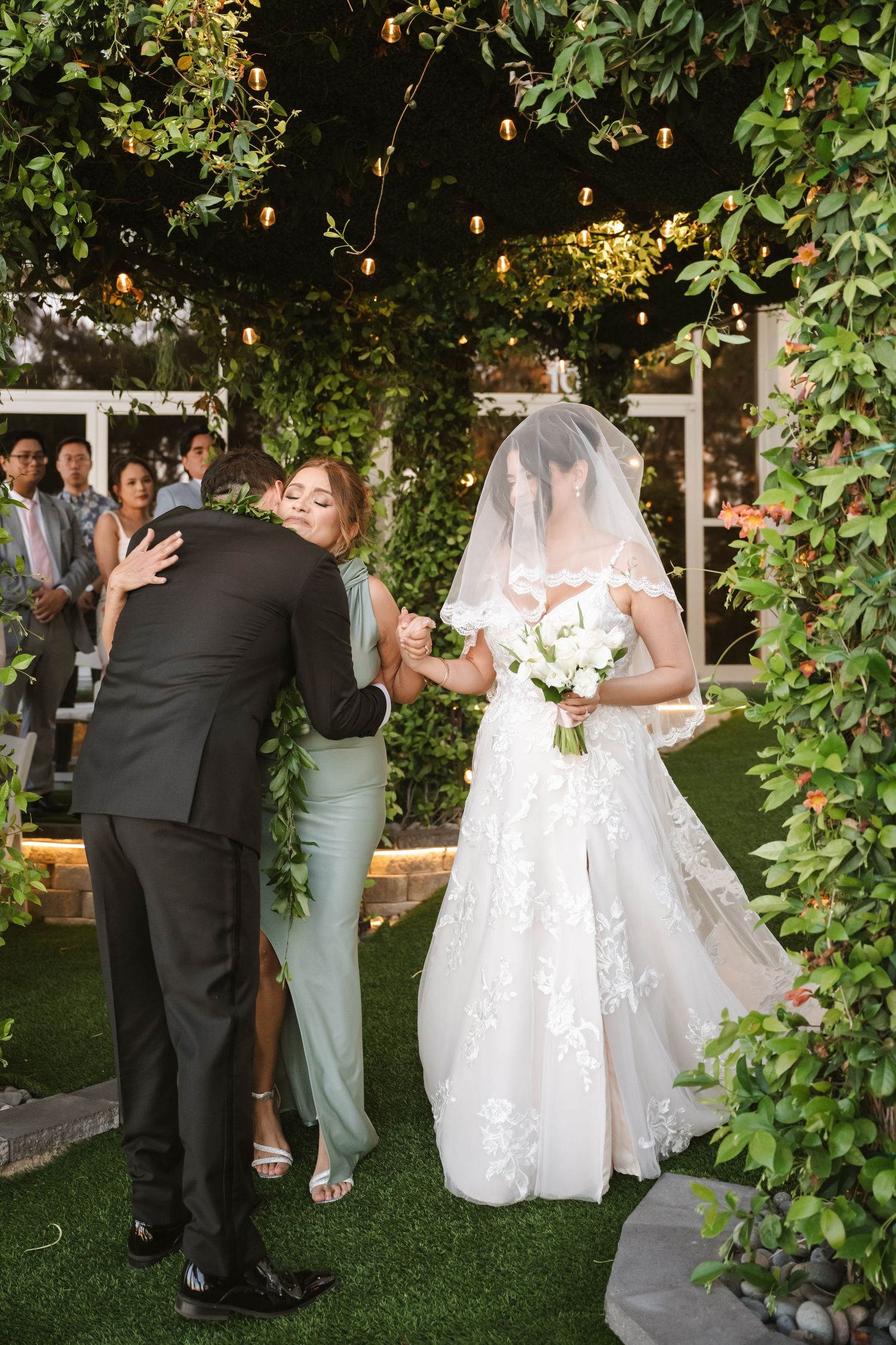 Groom embraces bridesmaid in sage dress as lace-gowned bride stands beside them at an elegant outdoor wedding ceremony.