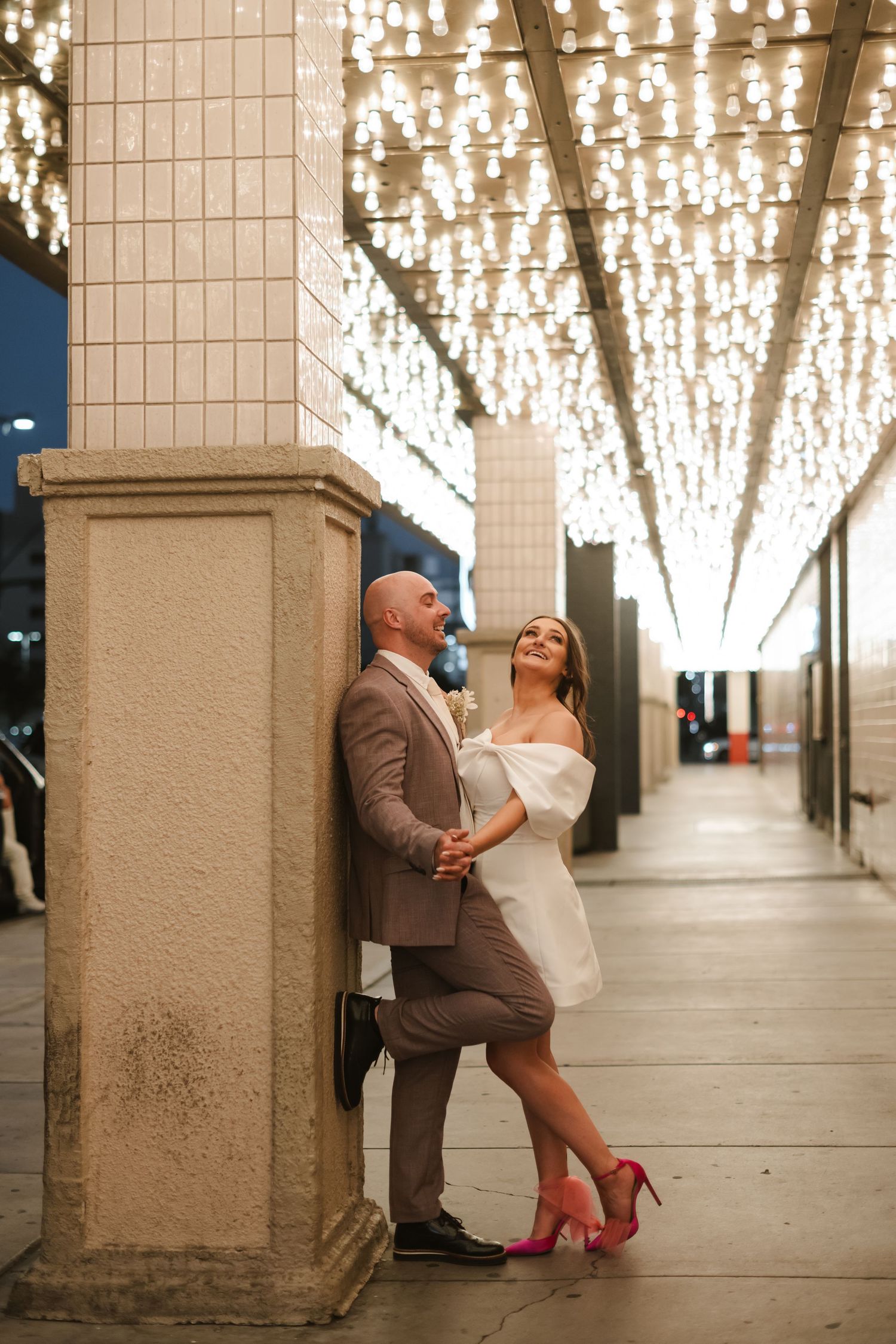 Couple in elegant attire embracing by a column under sparkling string lights in a romantic urban corridor.