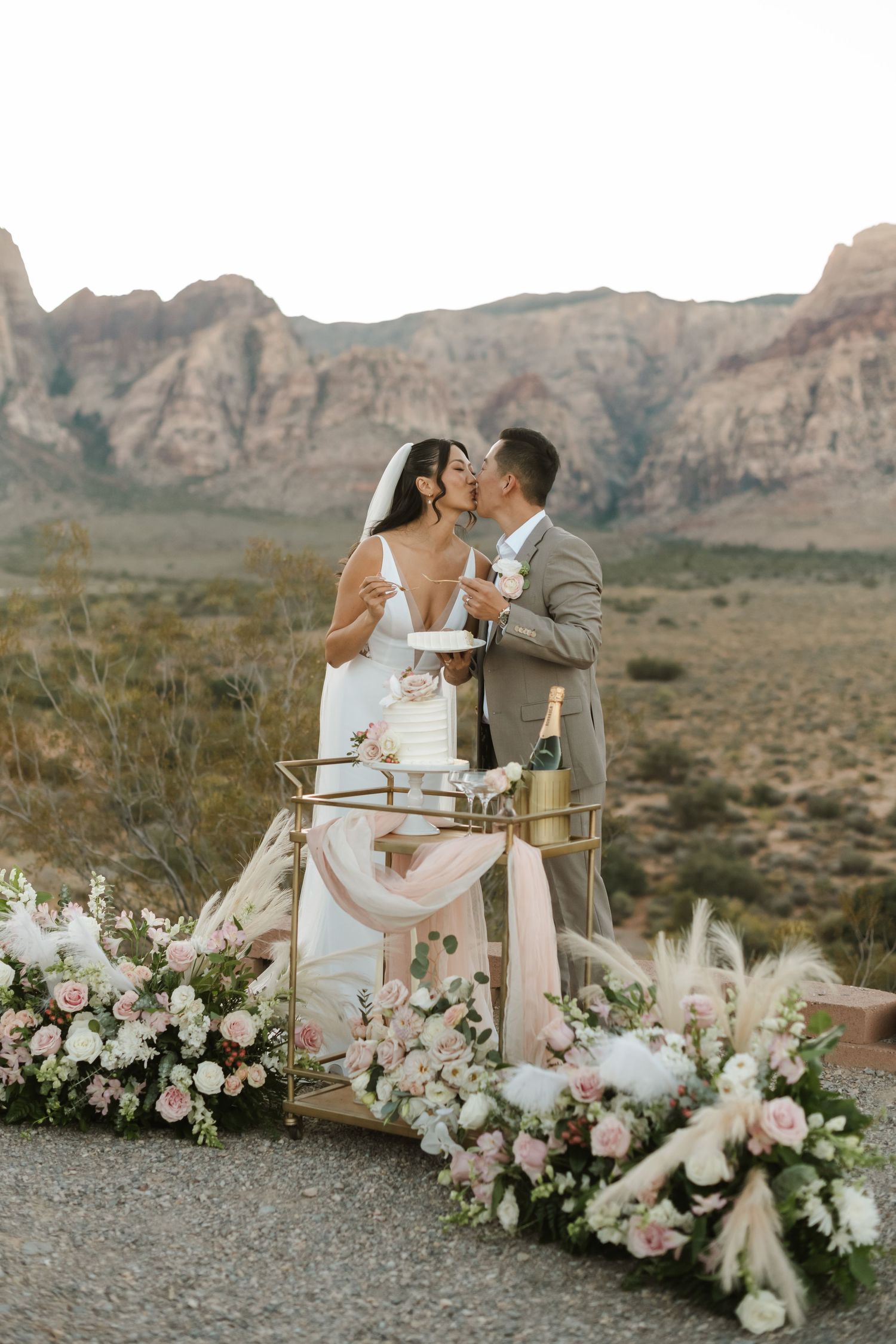 Couple kisses beside a wedding cake cart with pink floral arrangements, set against Red Rock Canyon desert landscape.