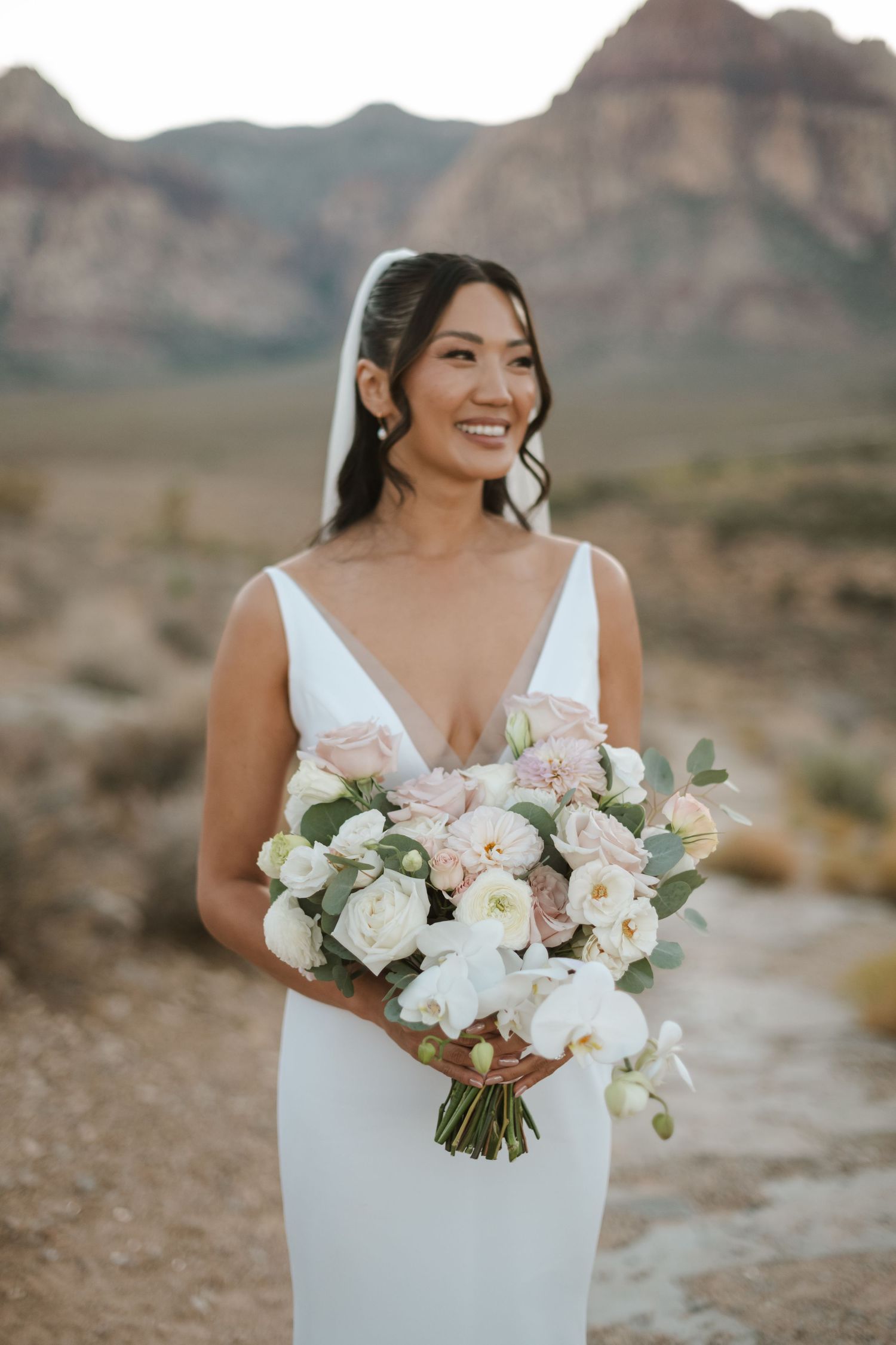 Smiling bride in white gown holds blush bouquet with desert mountains backdrop.