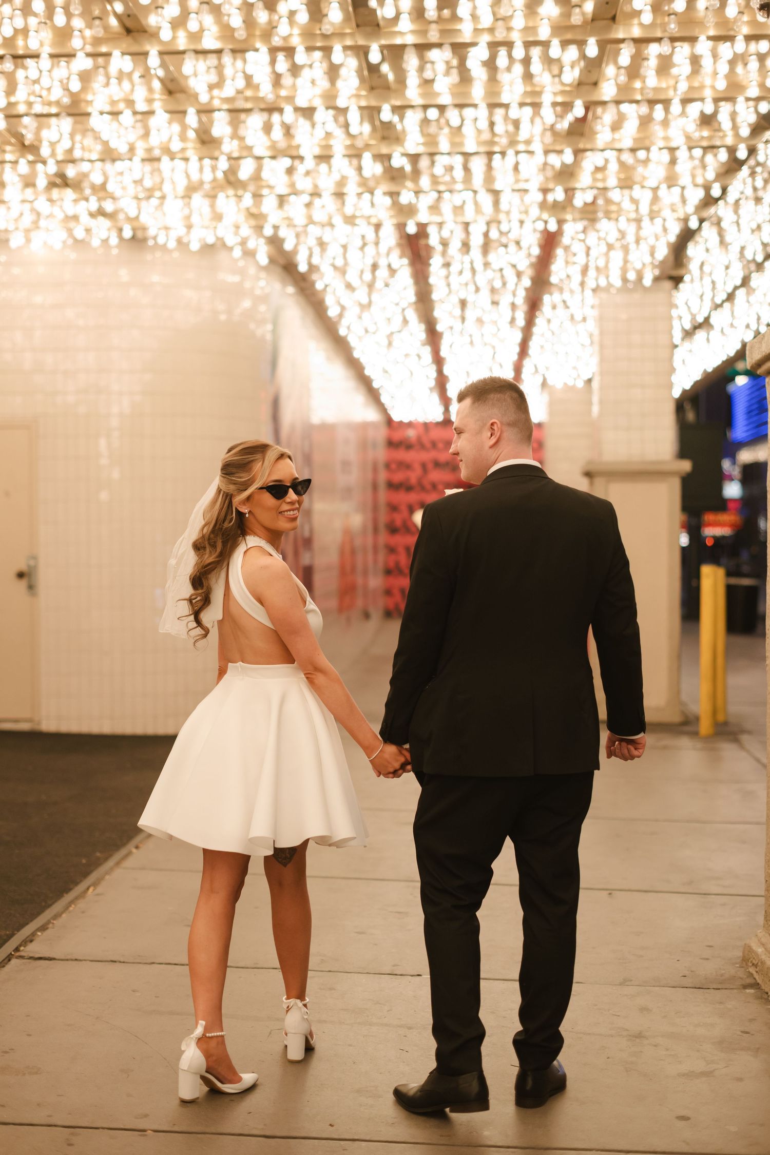 A couple walks under twinkling lights in an urban setting at night.