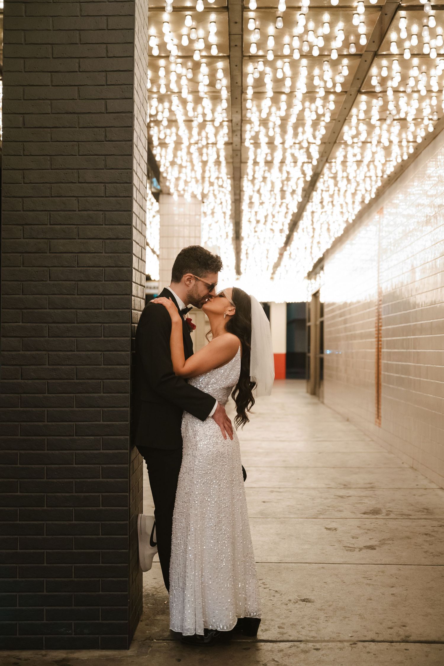 A romantic scene under twinkling lights in an industrial-style corridor with brick walls.