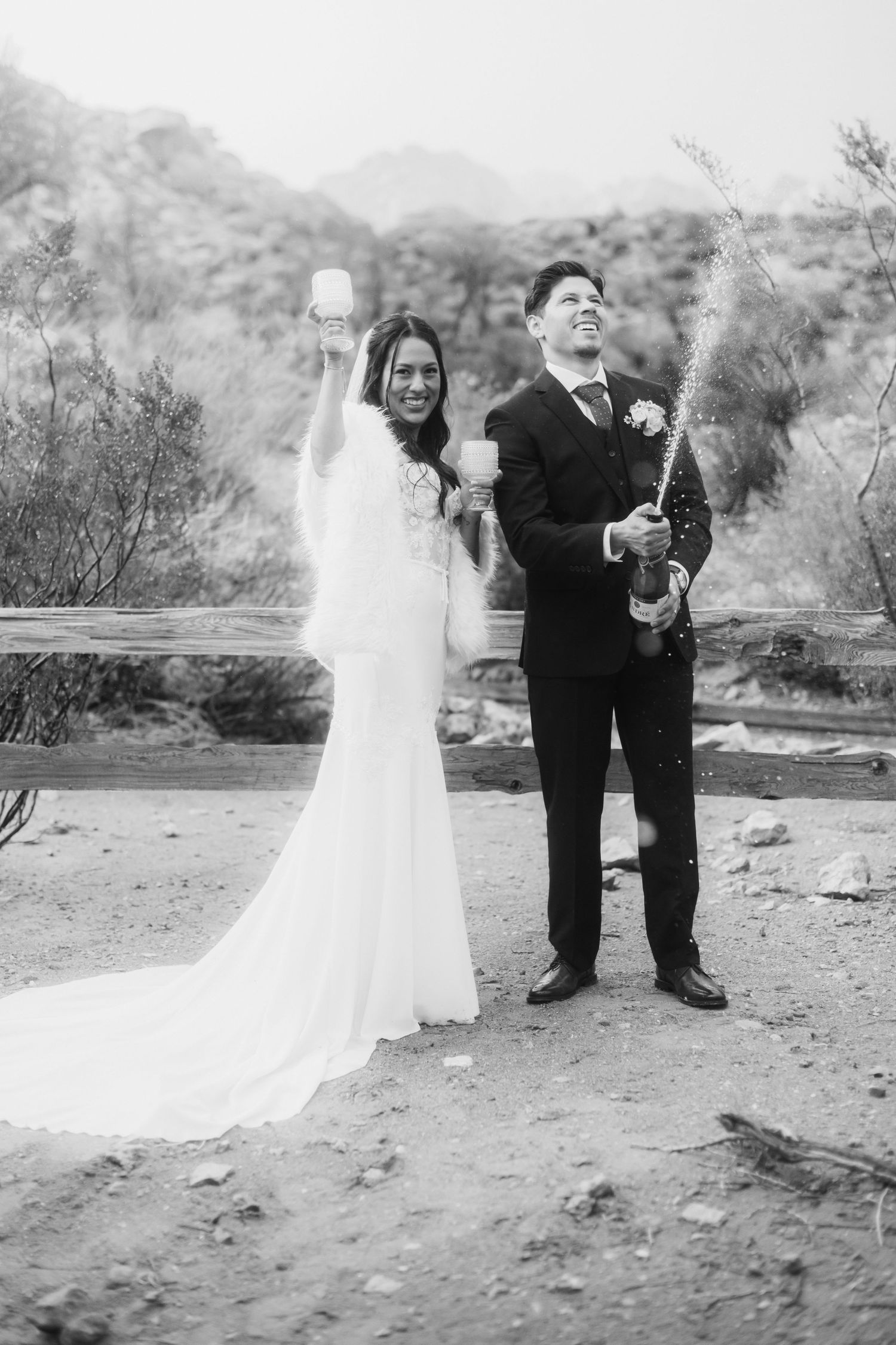 Black and white photo of a bride and groom celebrating their wedding by popping champagne outdoors in a desert setting.