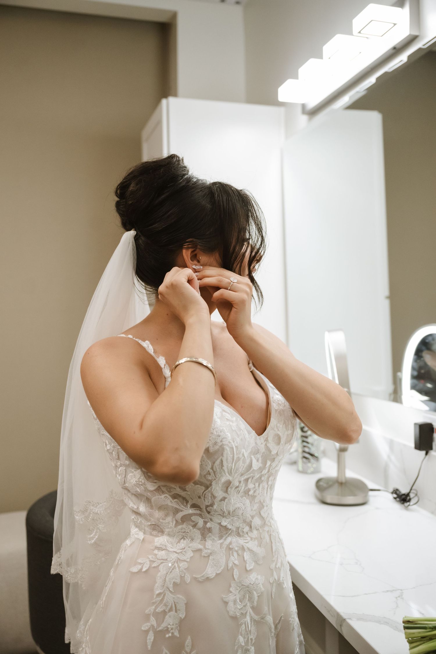 Bride in lace wedding dress and veil putting on earrings at vanity mirror while getting ready.