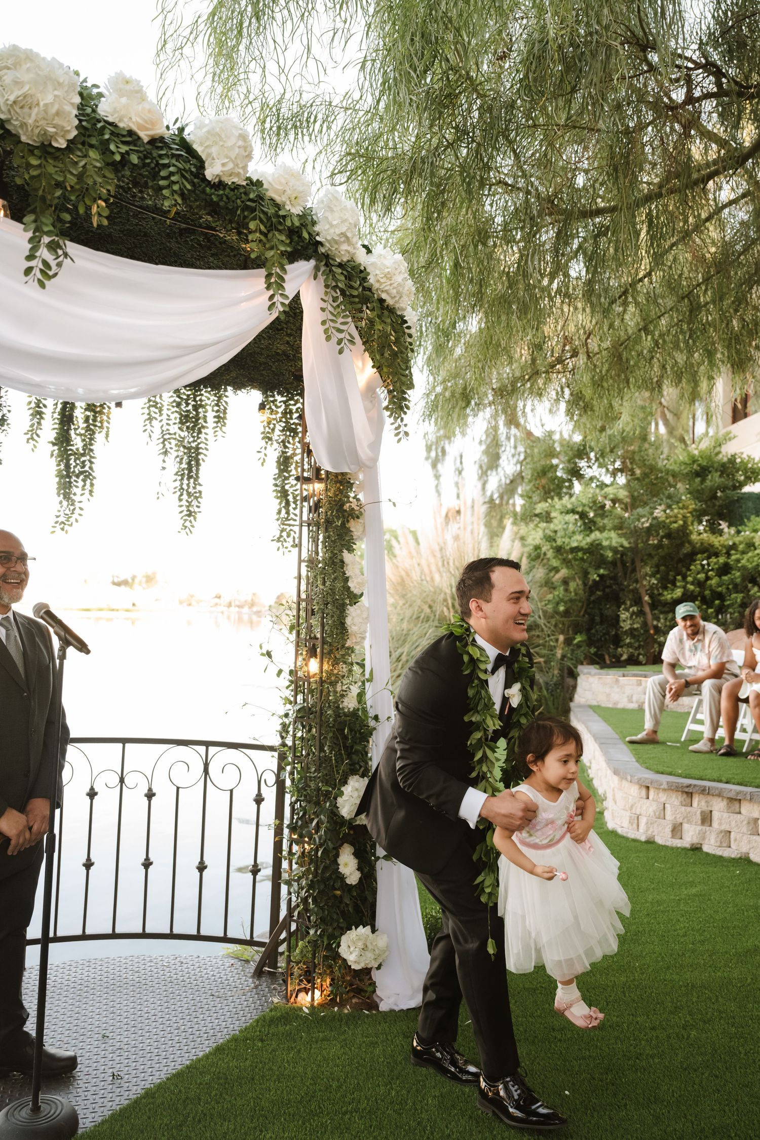 Groom in black suit laughs with flower girl in white dress at outdoor wedding ceremony under floral arch.