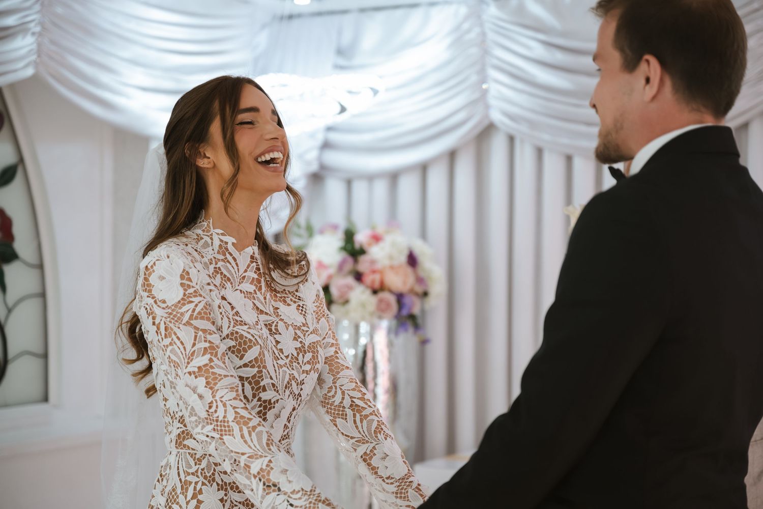 Bride in lace gown laughing joyfully with groom at wedding ceremony altar with floral decorations.
