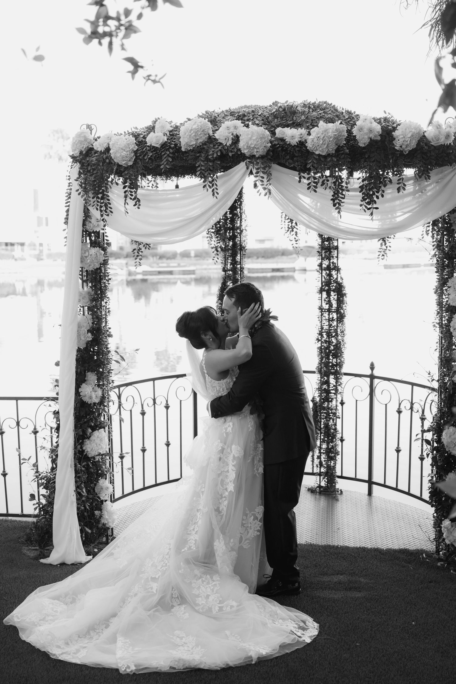 Black and white photo of bride and groom sharing first kiss under floral arch by waterfront.