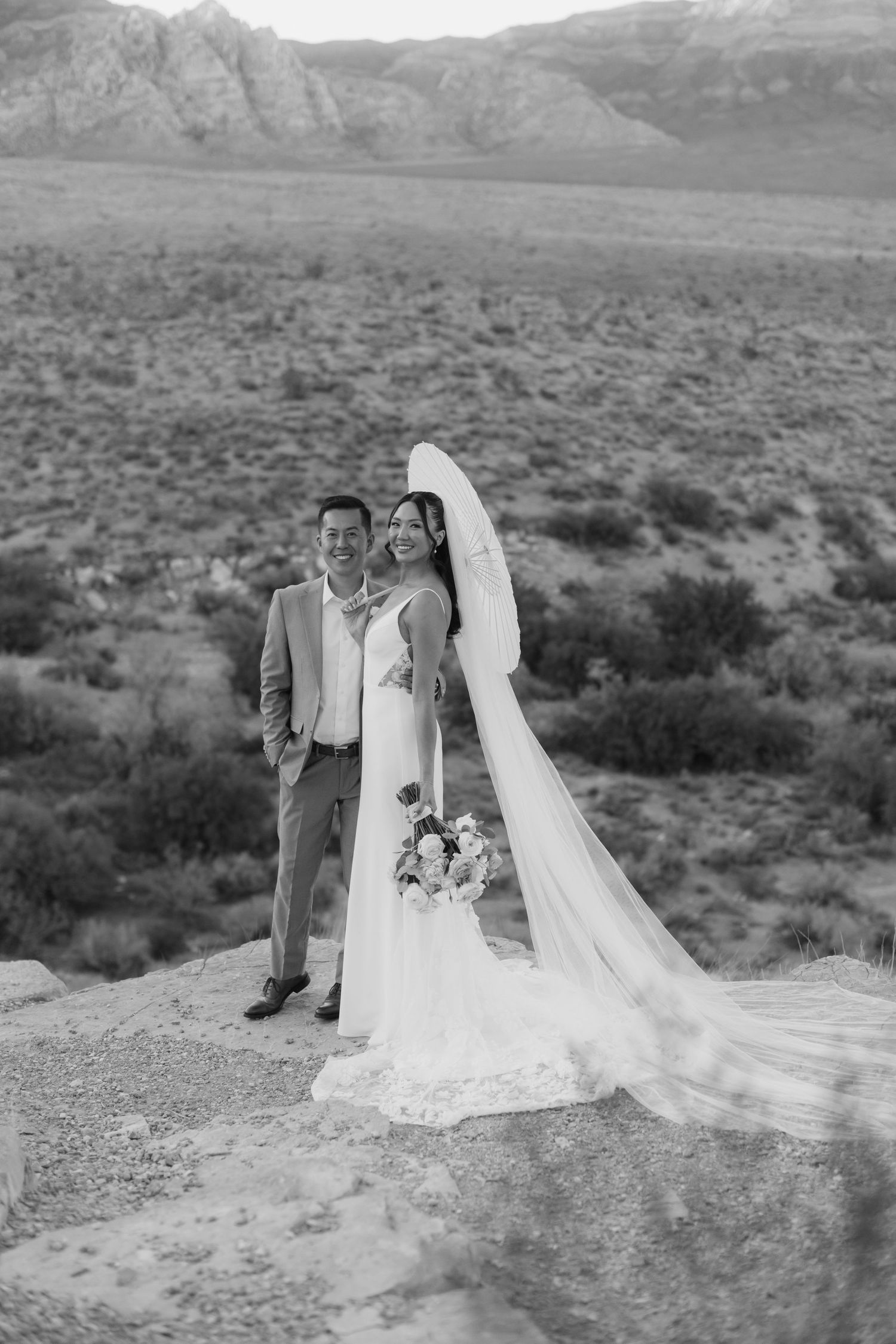 Black and white photo of a bride and groom posing on a rocky overlook with desert mountains in the background.