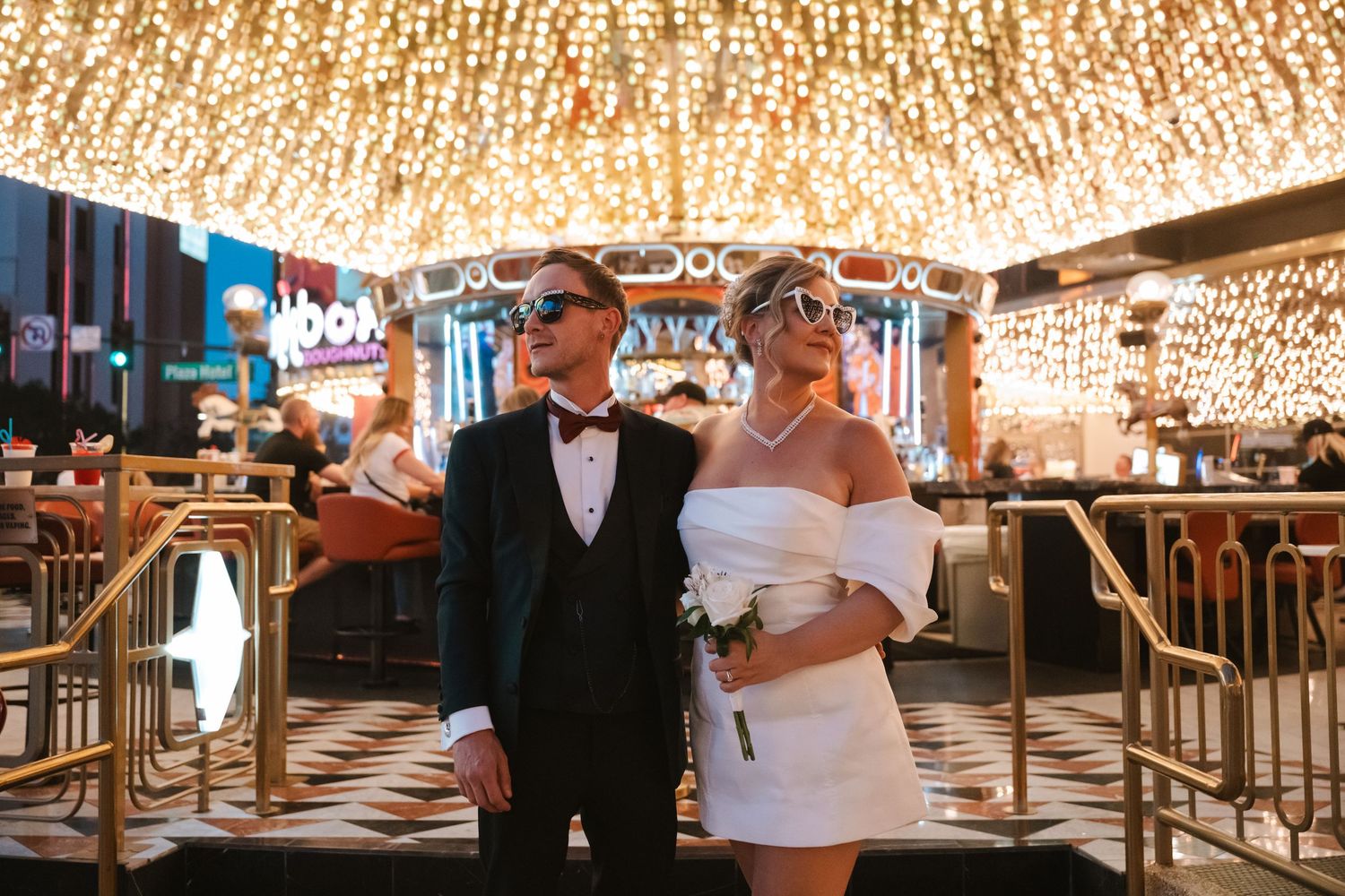 Newlywed couple in wedding attire wearing sunglasses pose under sparkling lights at a Las Vegas casino.