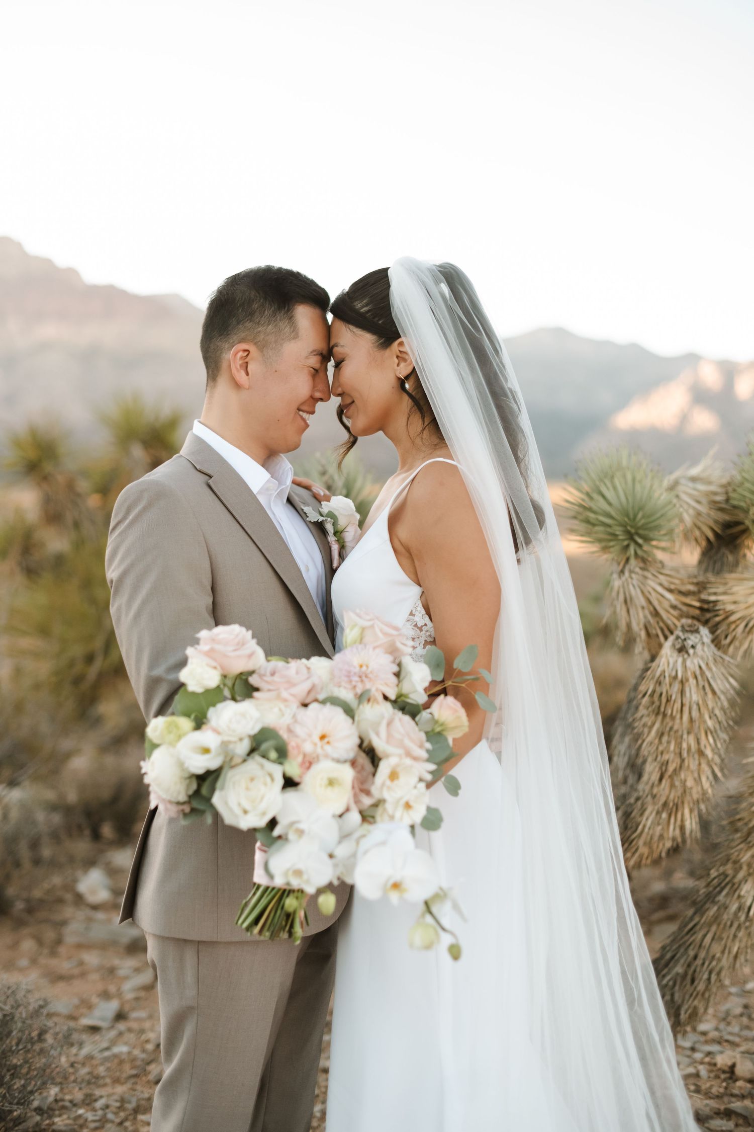 Bride and groom touch foreheads holding white and pink bouquet in desert landscape with mountains at golden hour.