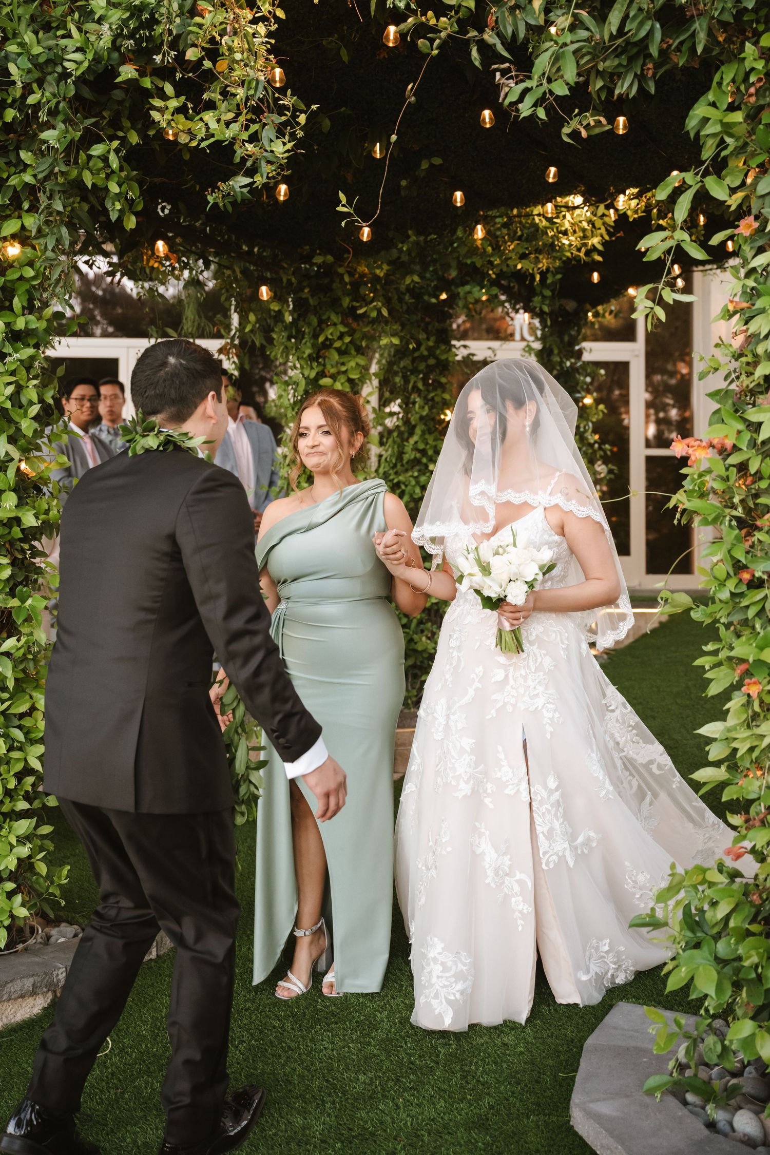 Groom in black suit meets bride in lace gown and veil, escorted by bridesmaid in sage dress at outdoor wedding.