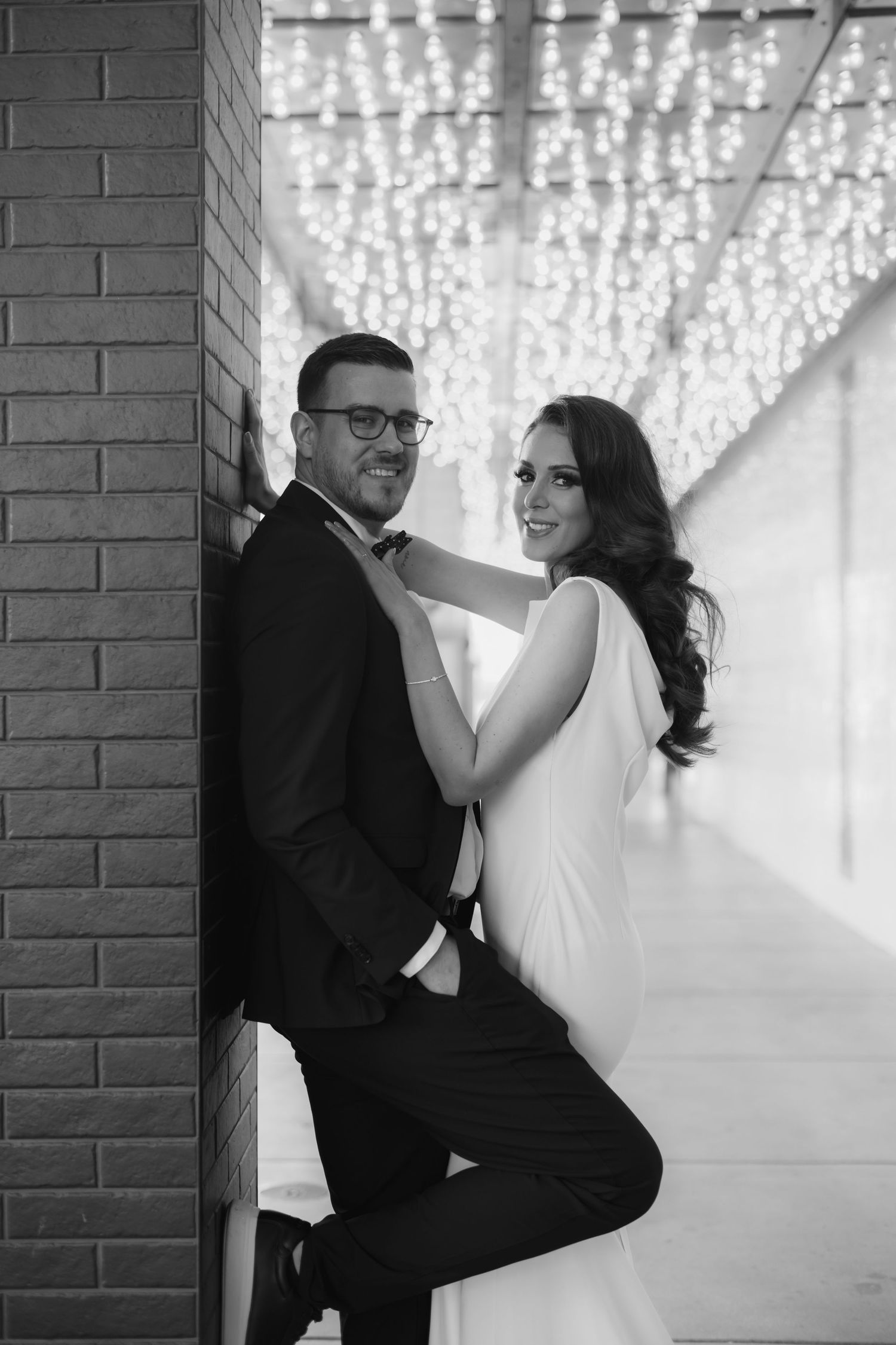 Black and white photo of a smiling couple in formal wear posing against a brick wall with bokeh lights.