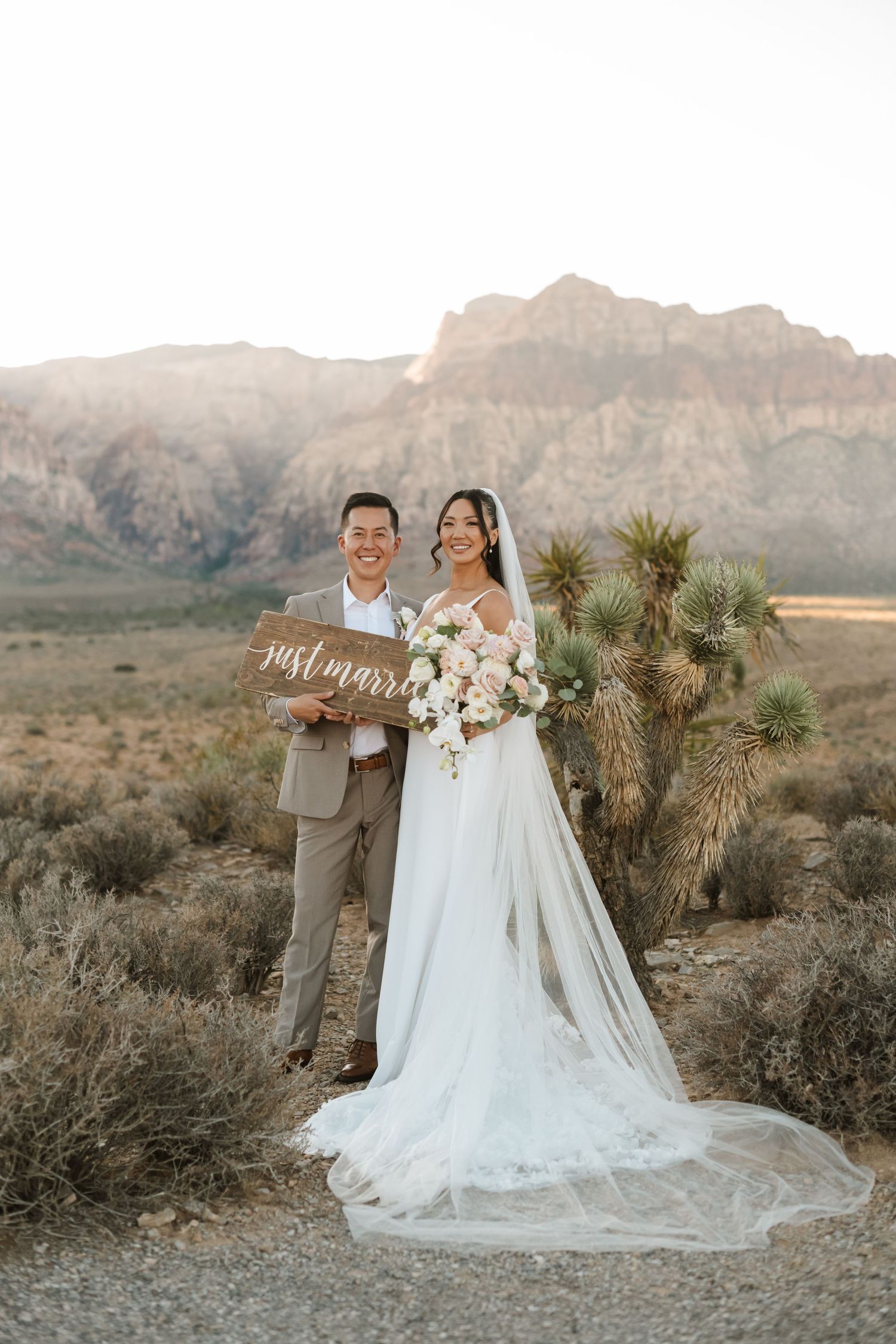 Newlyweds pose with Just Married sign at Red Rock Canyon desert wedding, bride in white gown with veil, groom in tan suit.