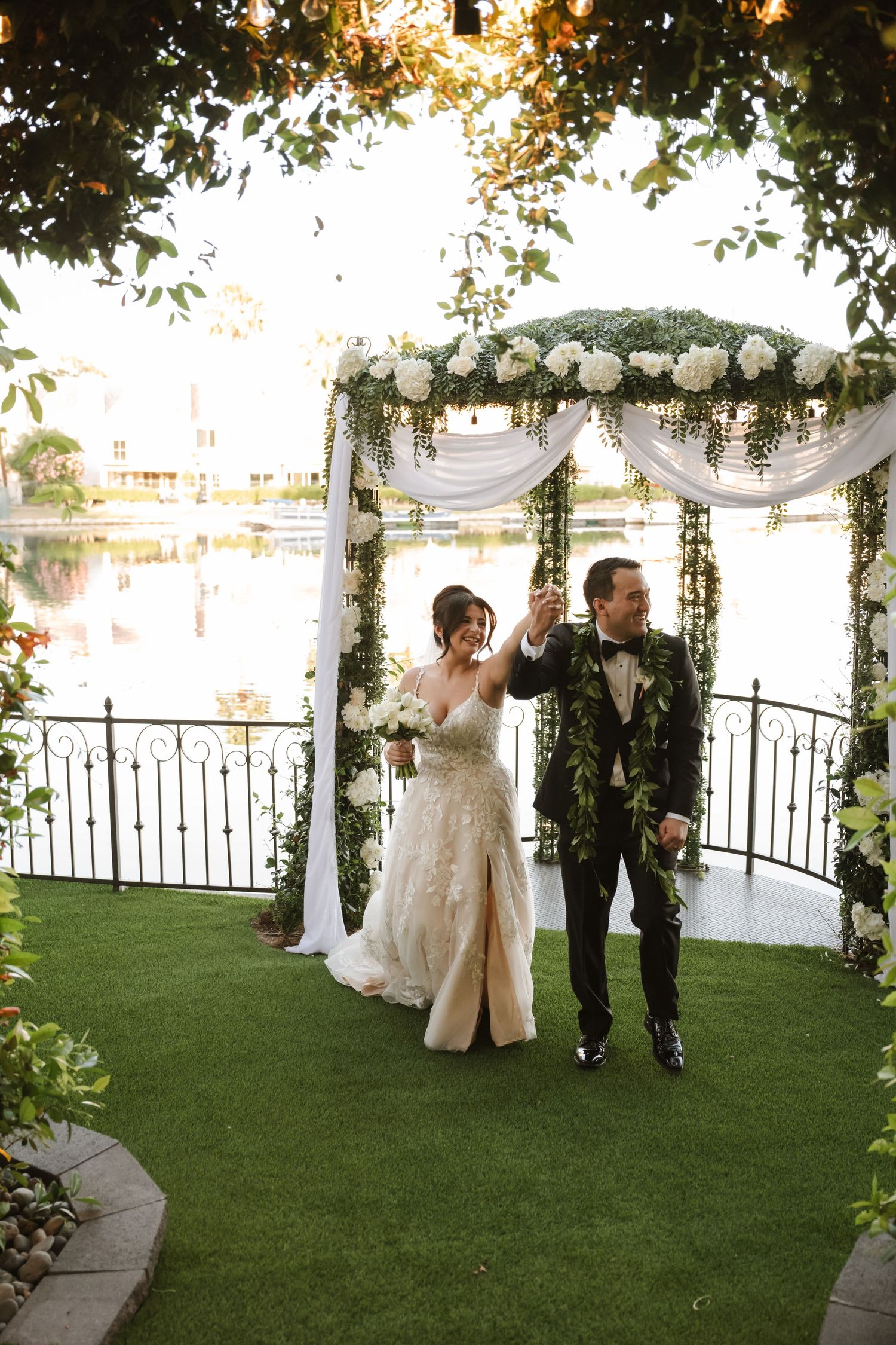 Bride and groom celebrate under floral arch with white draping by waterfront on lush green lawn.