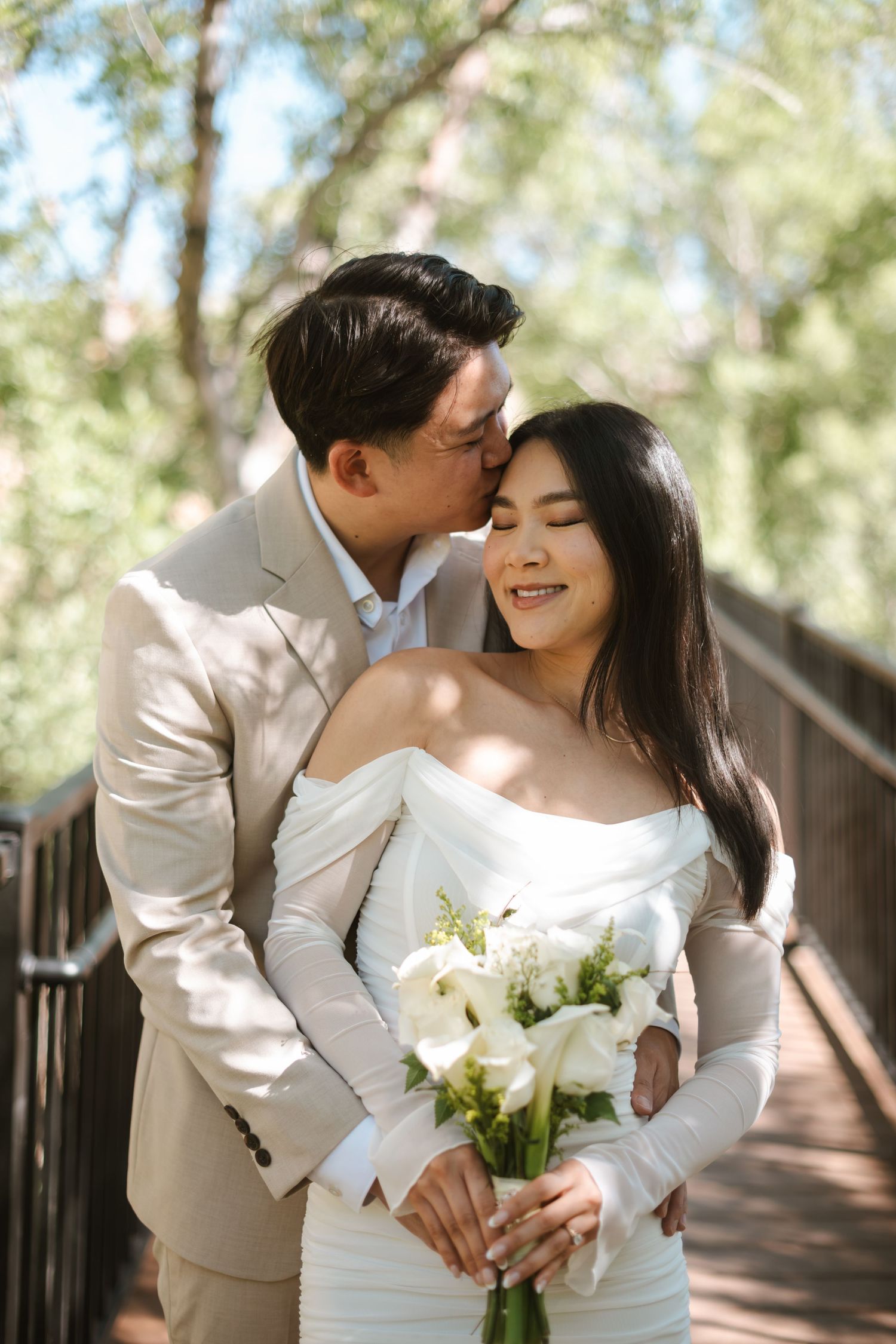 Groom in beige suit kissing bride on cheek as she smiles holding white calla lily bouquet on wooden bridge.