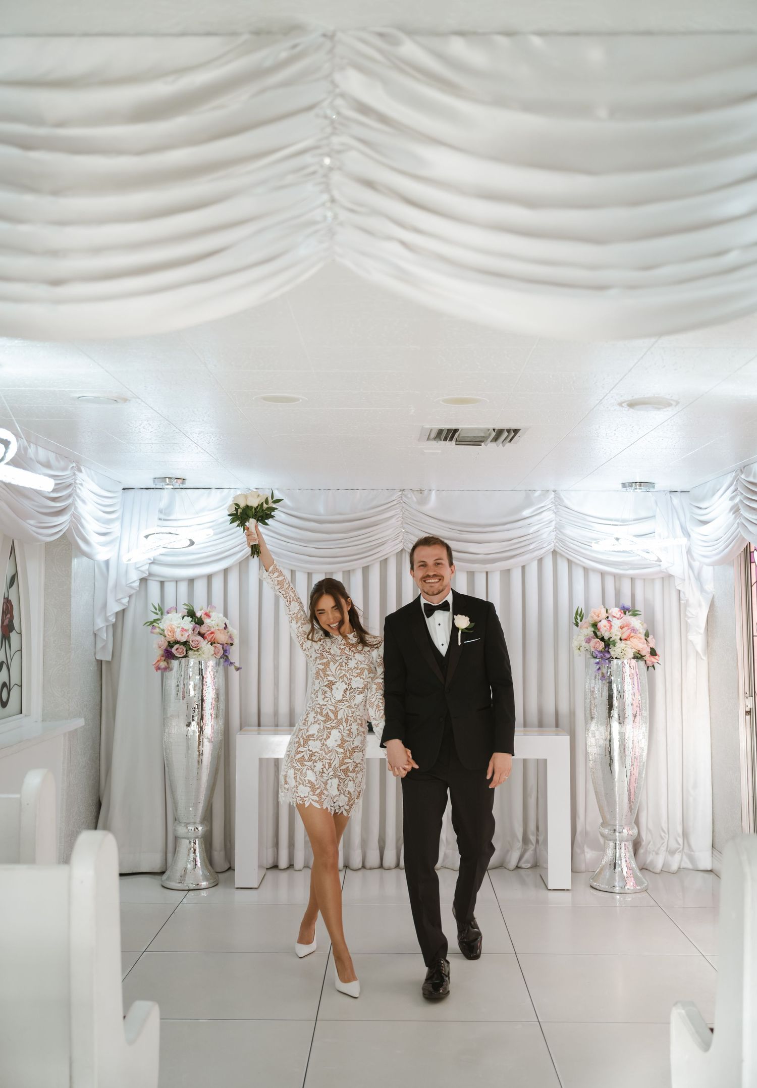 Joyful newlyweds celebrate in a white chapel, bride in short lace dress holding bouquet aloft, groom in black tuxedo.