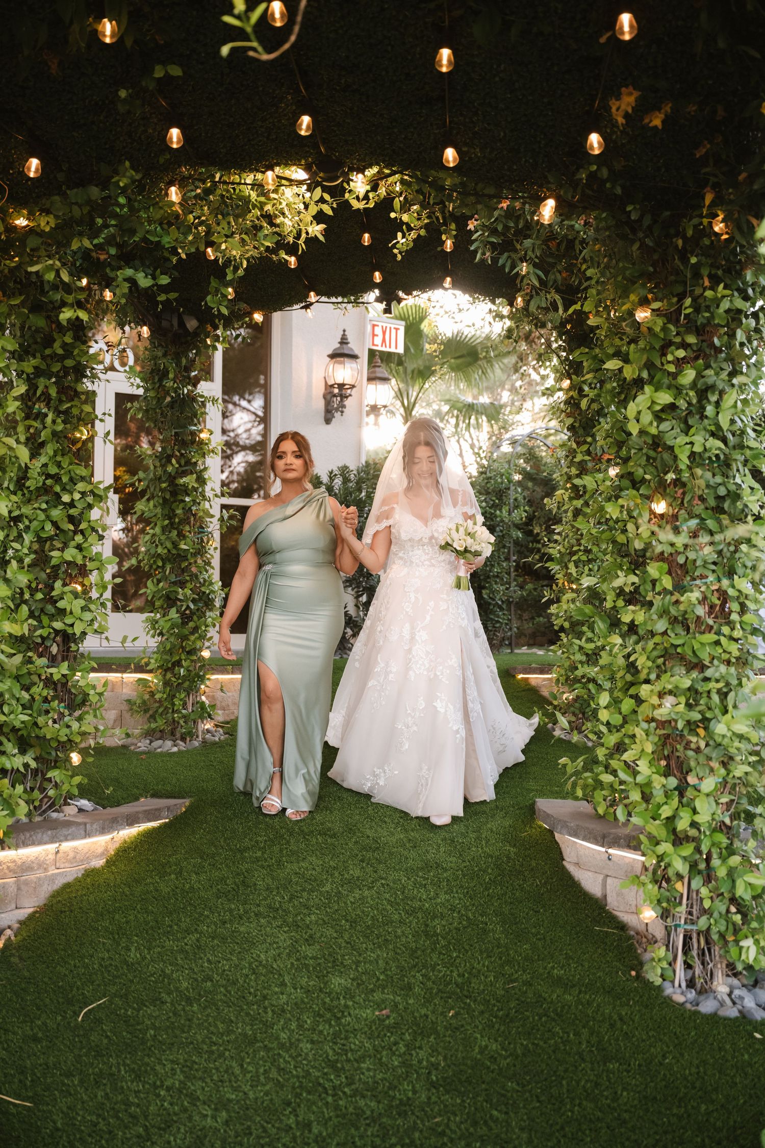 Bride in white lace gown walks with bridesmaid in sage green dress through ivy-covered archway with string lights.