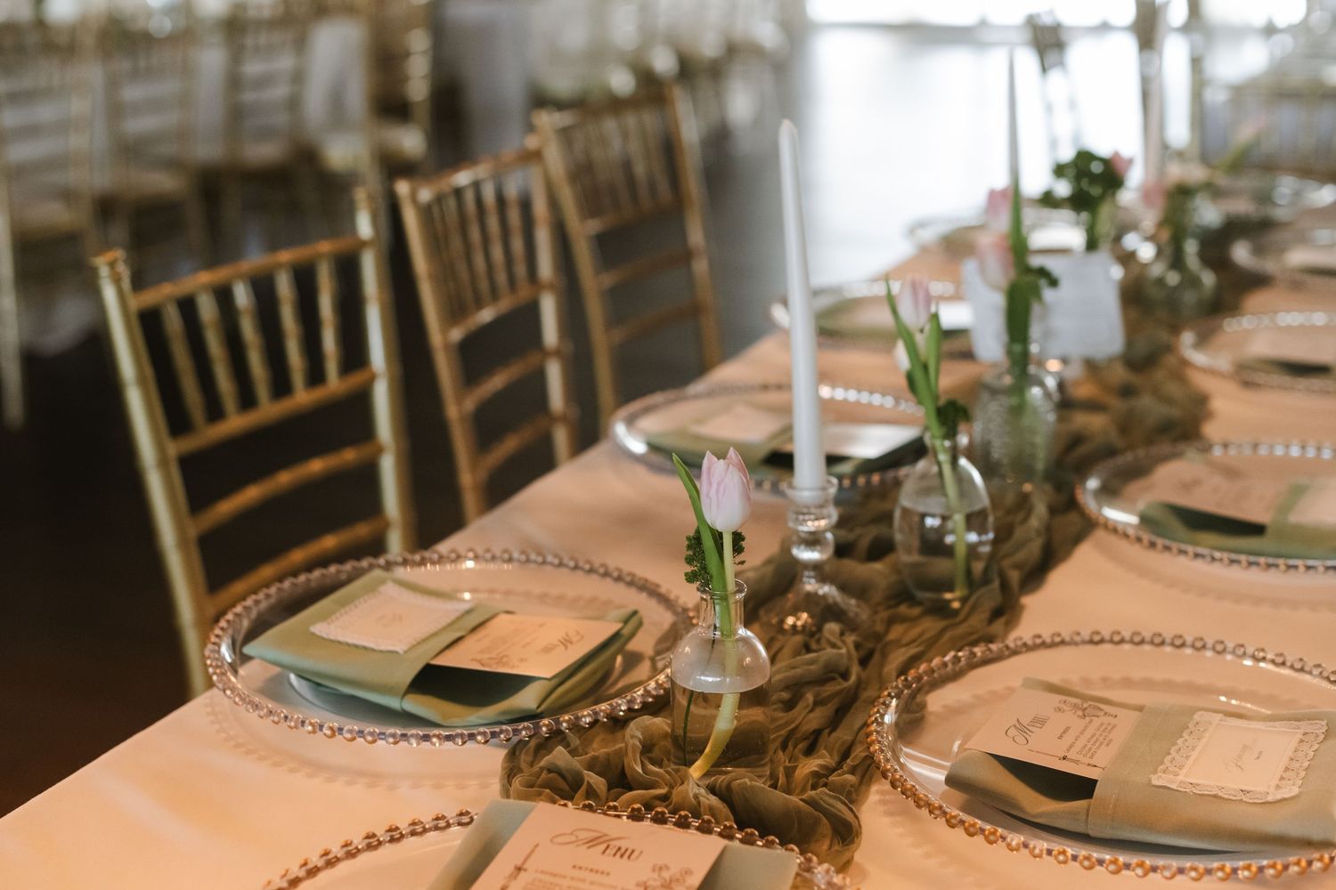 Elegant wedding reception table with gold chiavari chairs, beaded charger plates, taper candles, and a rustic burlap runner.