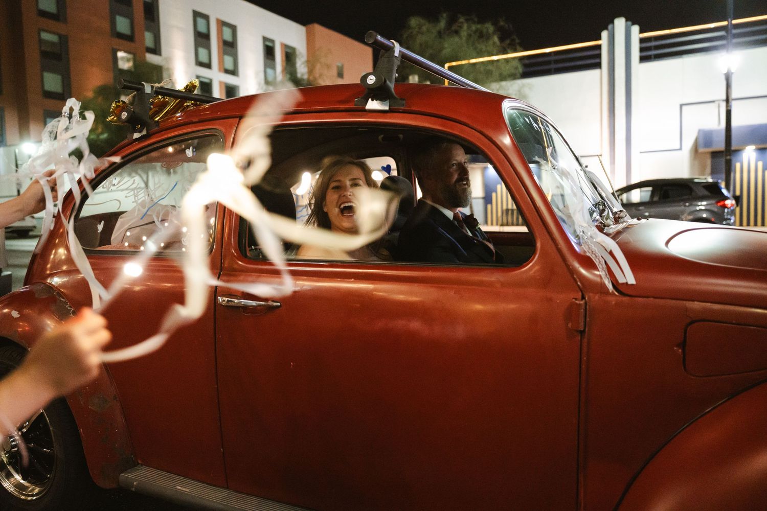 Bride and groom riding in a vintage red classic car decorated with wedding streamers at night.