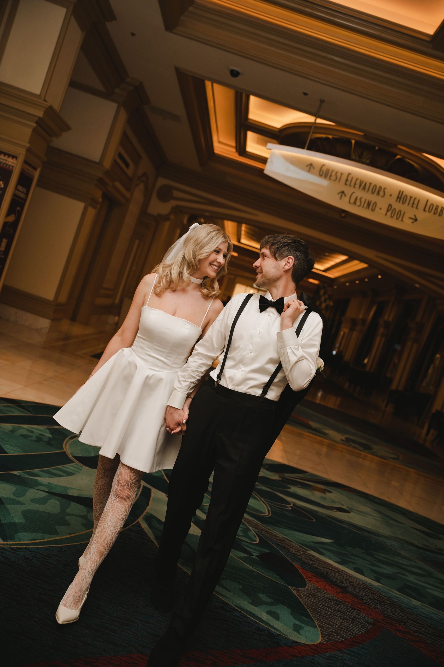 A bride in a short white dress and groom in black tuxedo with suspenders hold hands smiling in an elegant hotel lobby.