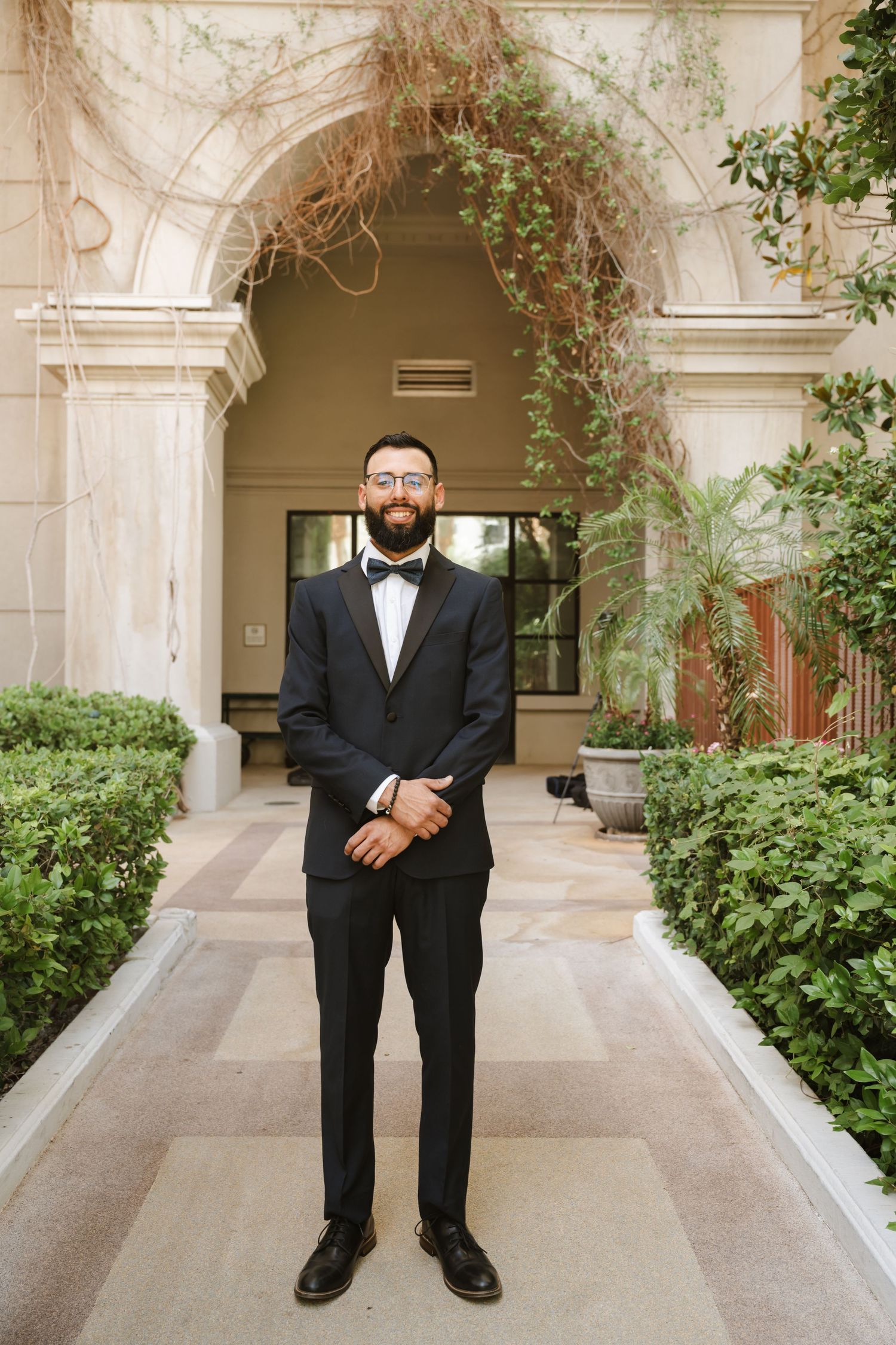 A groom in a classic black tuxedo poses in an elegant archway with climbing vines.