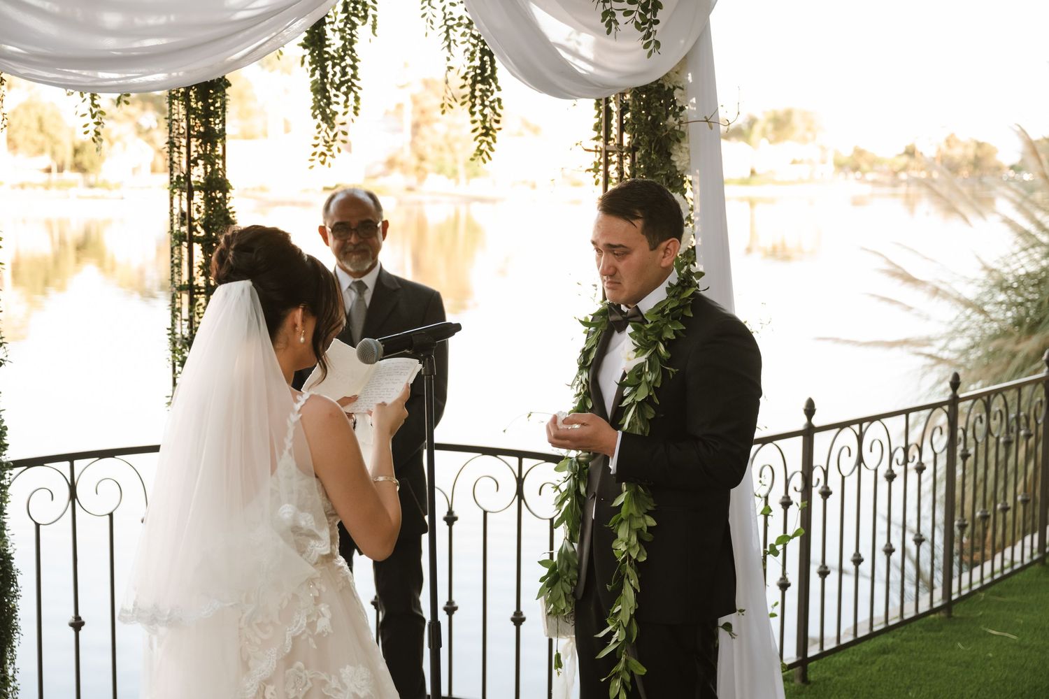 Groom wearing a green lei reads vows to bride under a decorated wedding arch near a serene lake.