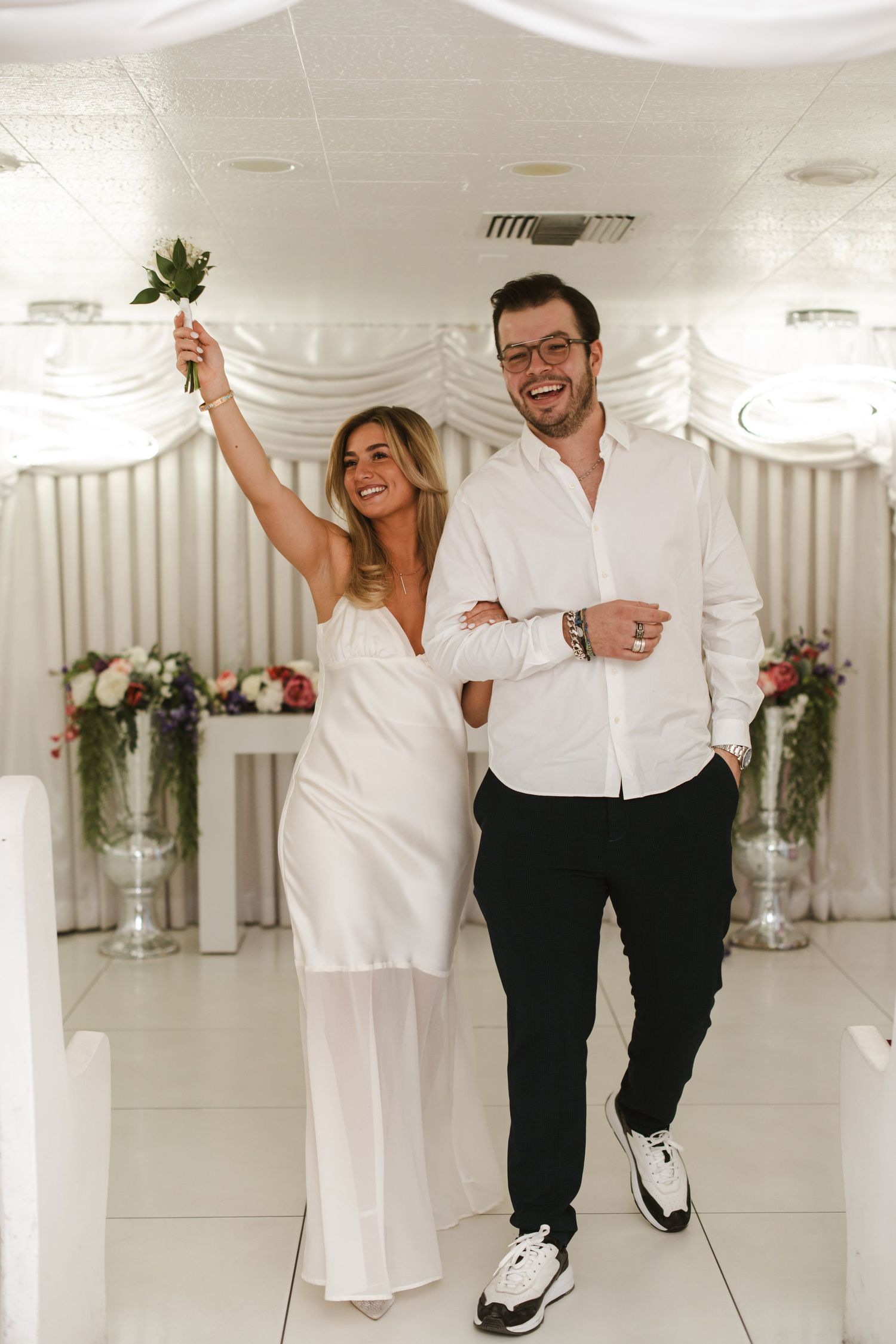 A couple dances and laughs together in a bright white wedding venue with floral arrangements.