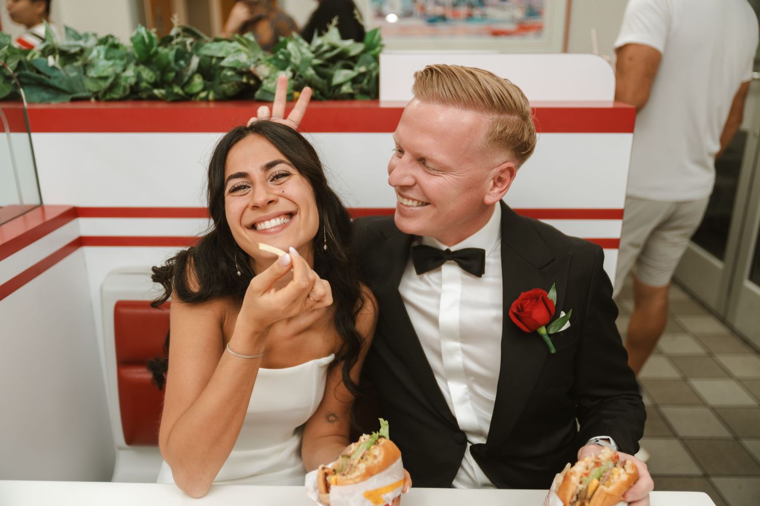 Bride and groom laughing together at a diner booth, enjoying hot dogs in wedding attire with a red rose boutonniere.