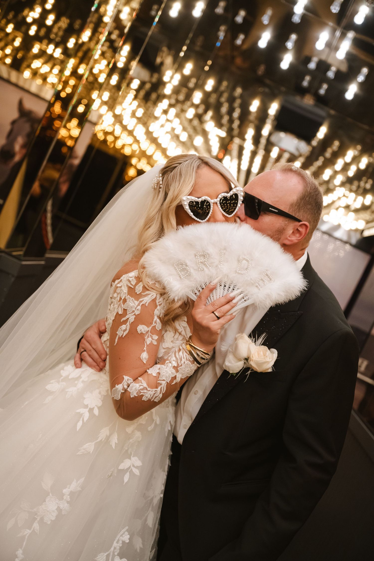 Bride and groom kissing behind a white feather fan, wearing stylish sunglasses, with warm bokeh lights in the background.