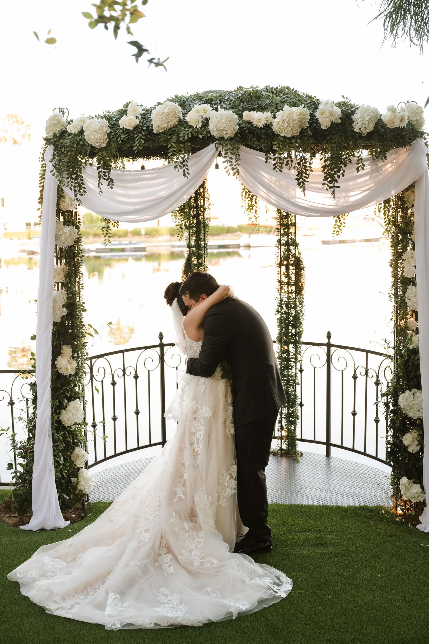 Bride and groom share first kiss under floral arch adorned with white roses by lake.